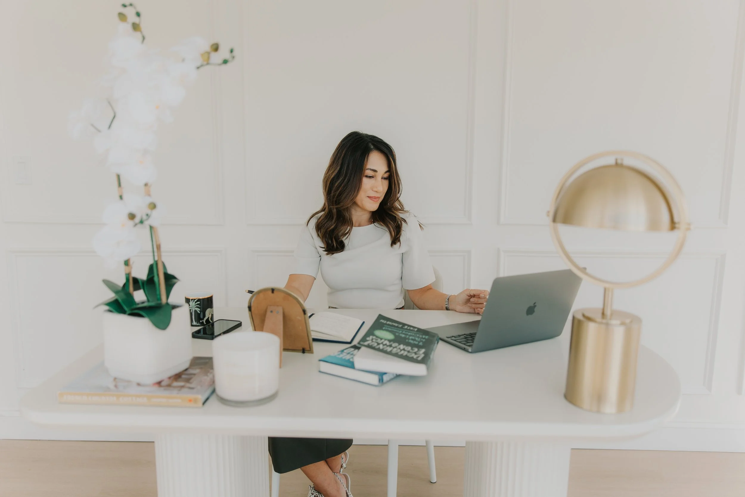 Woman sitting at a white desk with a silver laptop, open books, a pen holder, and decorative items including a golden lamp and a white orchid in a pot.