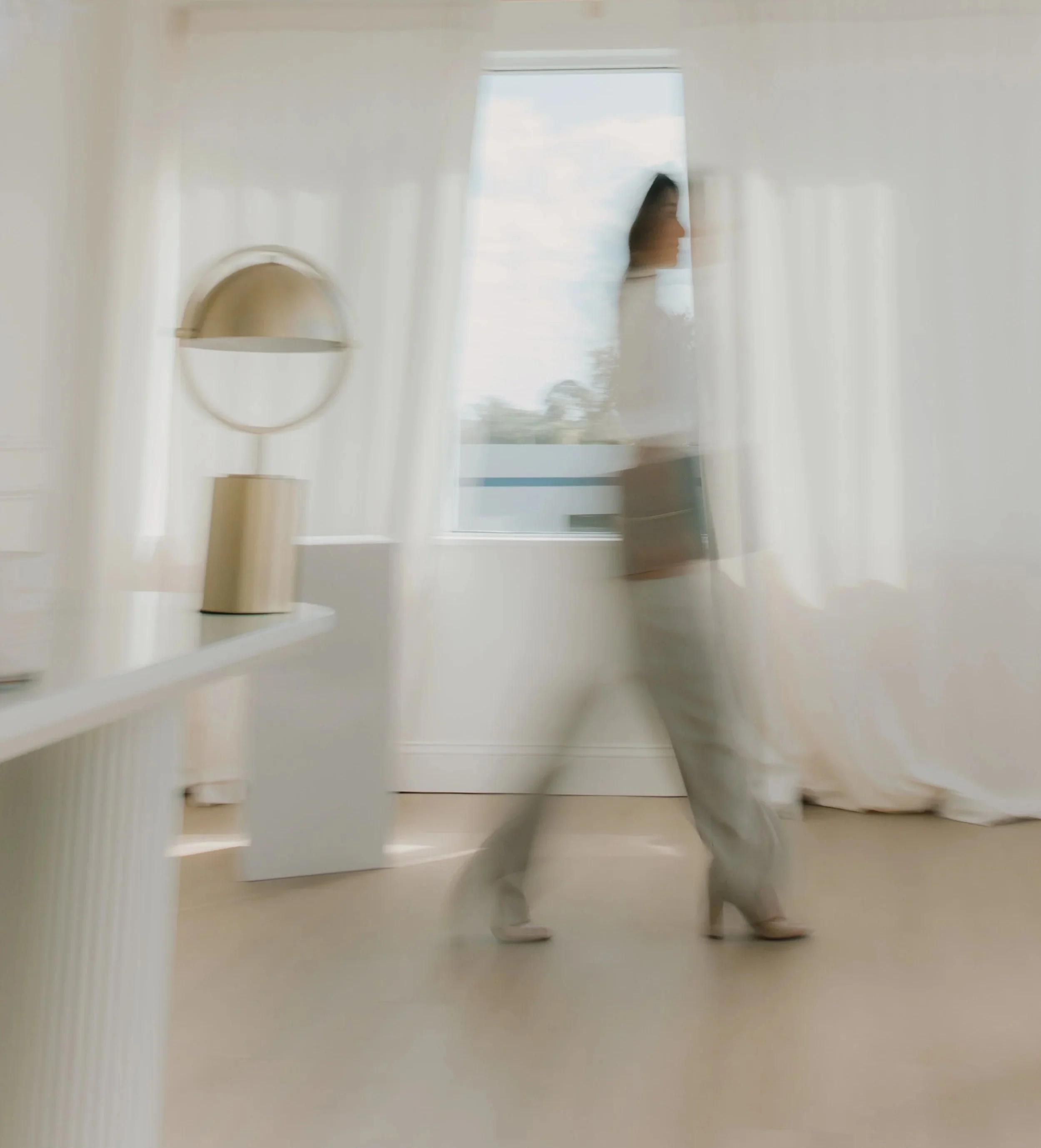 A person walking through a bright room with white curtains, a table lamp, and a window showing a cloudy sky.