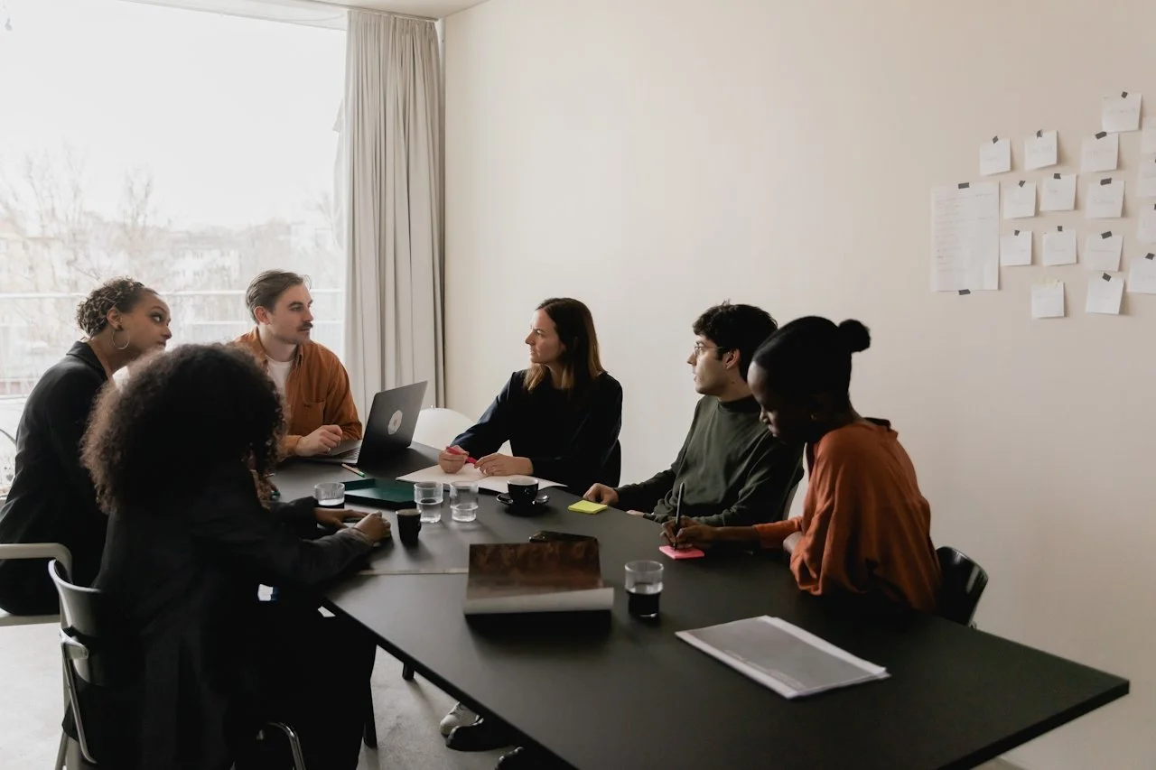 Six people gathered around a black conference table in a well-lit room with a large window and white curtains. They are engaged in a meeting, with some taking notes and others listening.