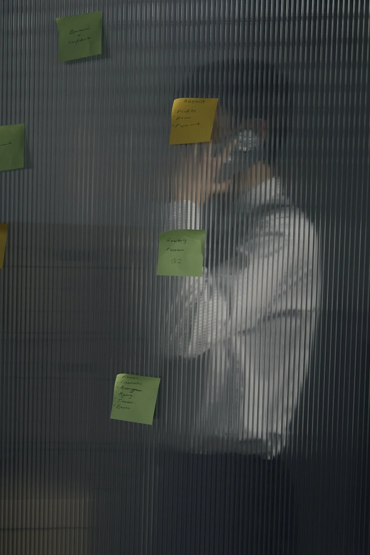Person taking a photo of a glass wall with sticky notes attached, including a yellow sticky note labeled 'AGENDA' and green sticky notes with handwritten text.