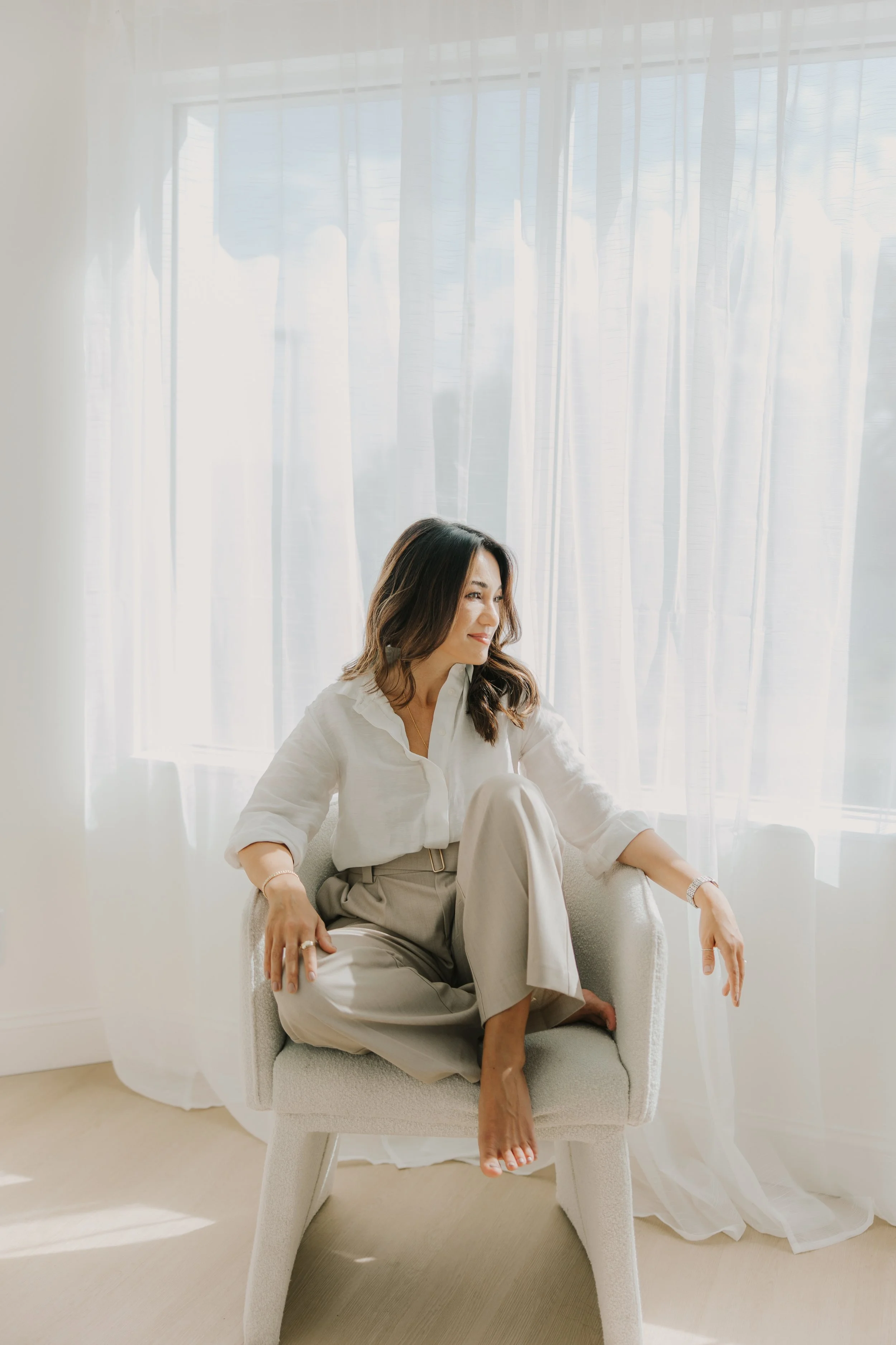 A woman with brown hair wearing a white blouse and beige pants sitting in a white armchair near a large window with sheer white curtains, with sunlight streaming in.