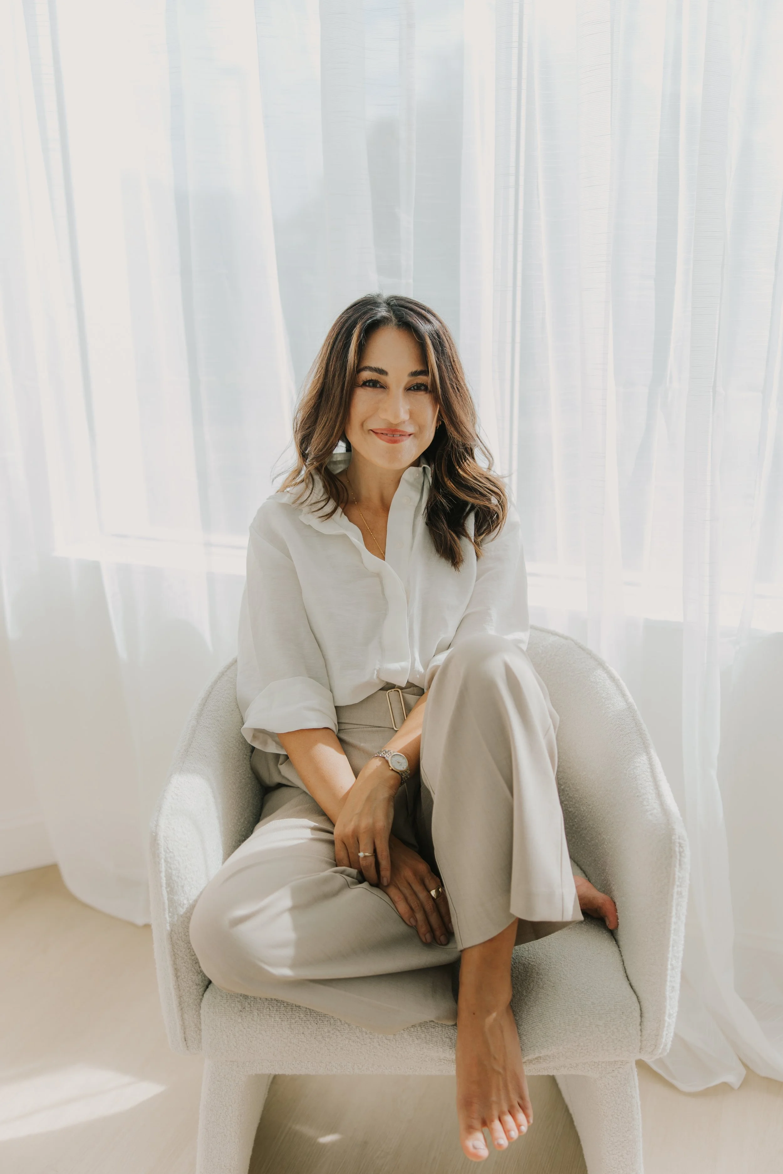 A woman with shoulder-length brown hair, wearing a white blouse and beige pants, sitting on a white chair in front of sheer white curtains and smiling at the camera.