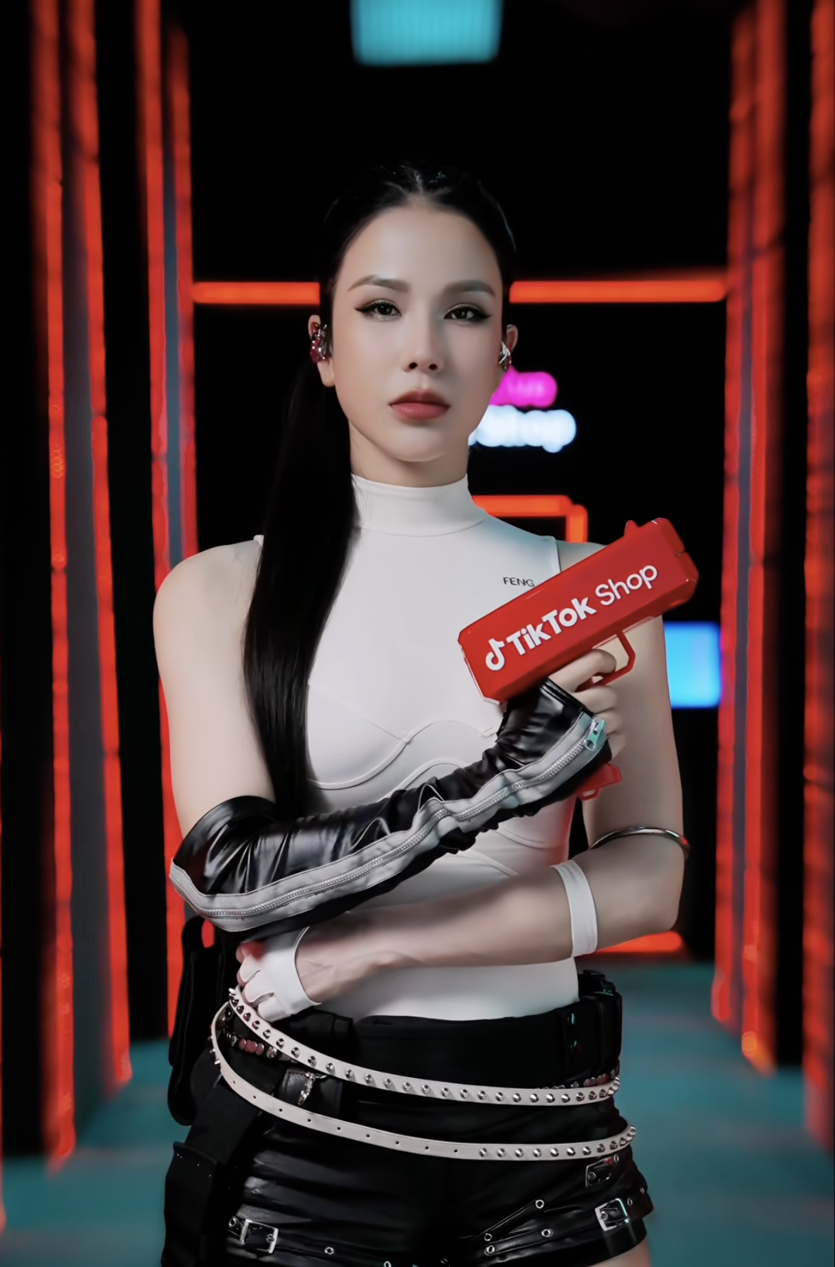 A woman with long black hair, dressed in a futuristic white top and black leather pants, holds a red TicTok Shop sign in a cyber-themed setting with neon lights.