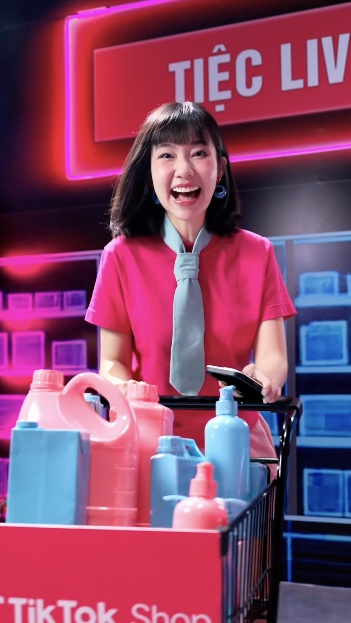 Young woman smiling and looking at her phone while standing behind a shopping cart filled with colorful bottles of laundry detergent in a TikTok Shop setting, with vibrant pink and blue neon signage in the background.
