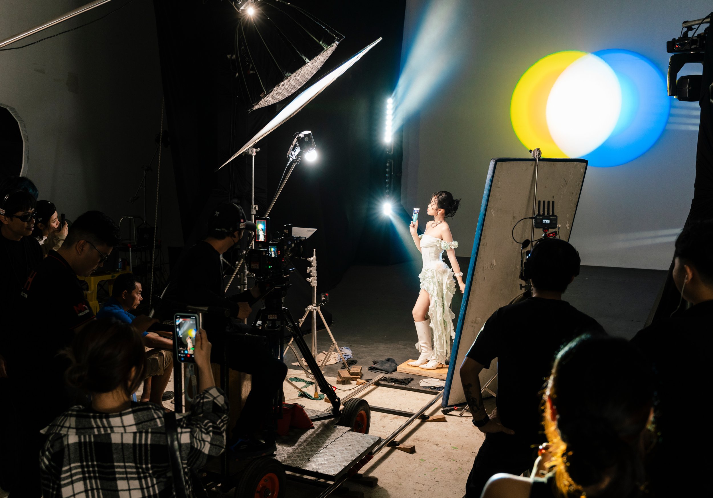 A photoshoot studio with a model posing in left profile, wearing a ruffled white dress, knee-high boots, and holding a phone. Multiple crew members operate cameras and lighting, with various equipment and reflectors in the scene.