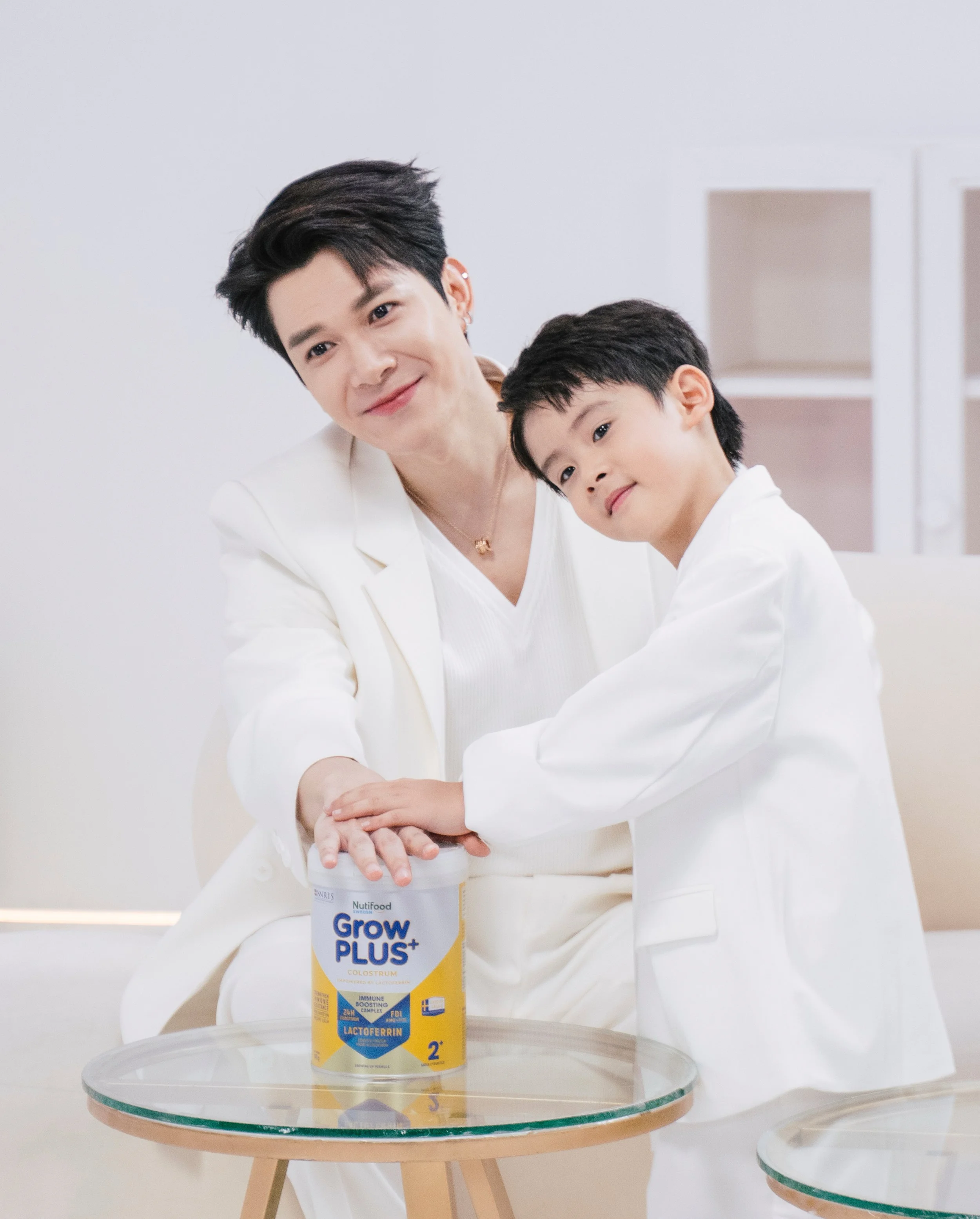A woman and a young boy, both dressed in white, are smiling and holding a container of Nutifood Grow Plus+ on a glass table. The woman is seated while the boy stands beside her, both looking at the camera.
