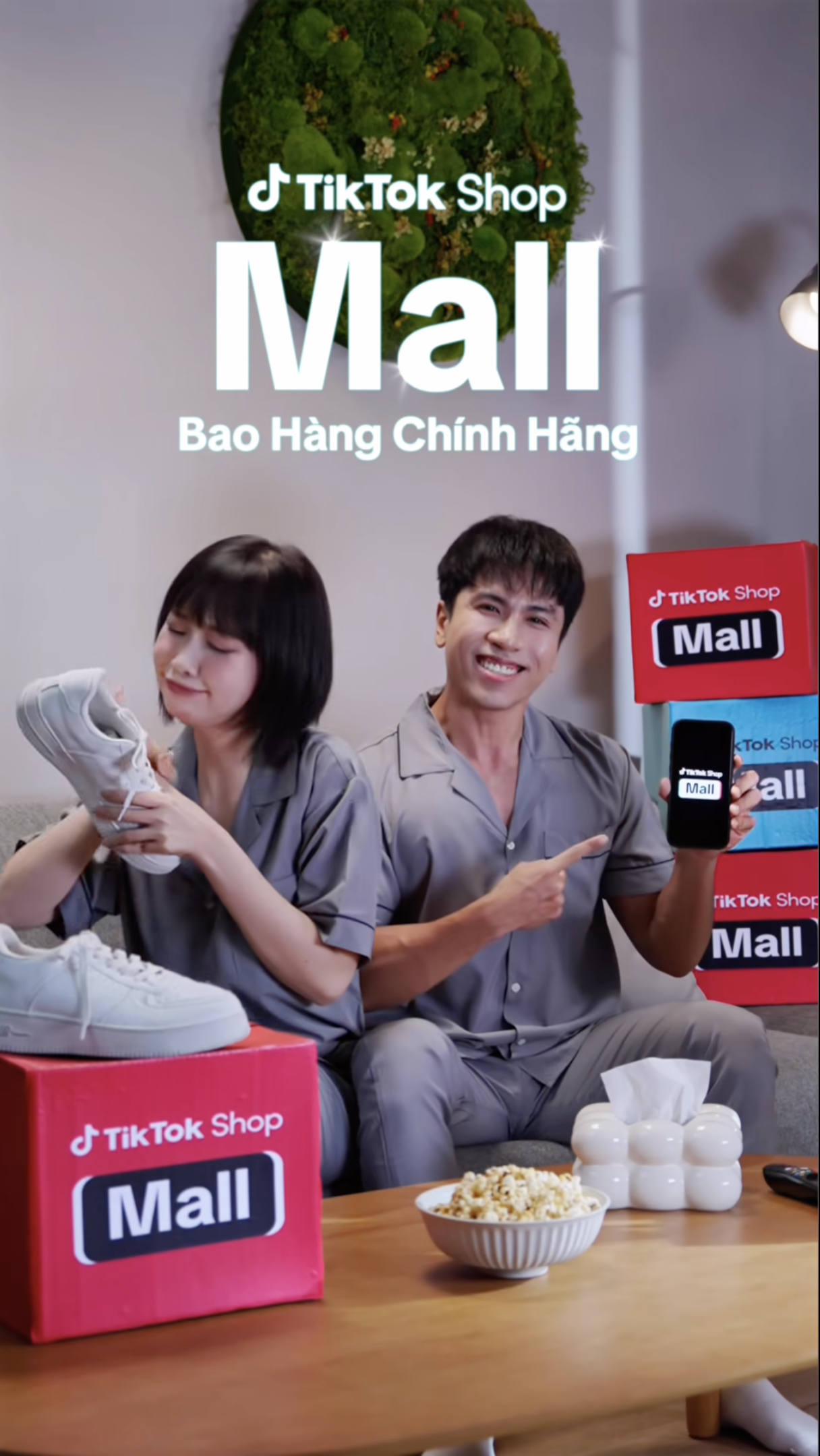 Two people sitting on a couch, holding products and pointing at them, with TikTok Shop Mall boxes around them, a bowl of popcorn on the table, and a tissue box, promoting TikTok Shop Mall.
