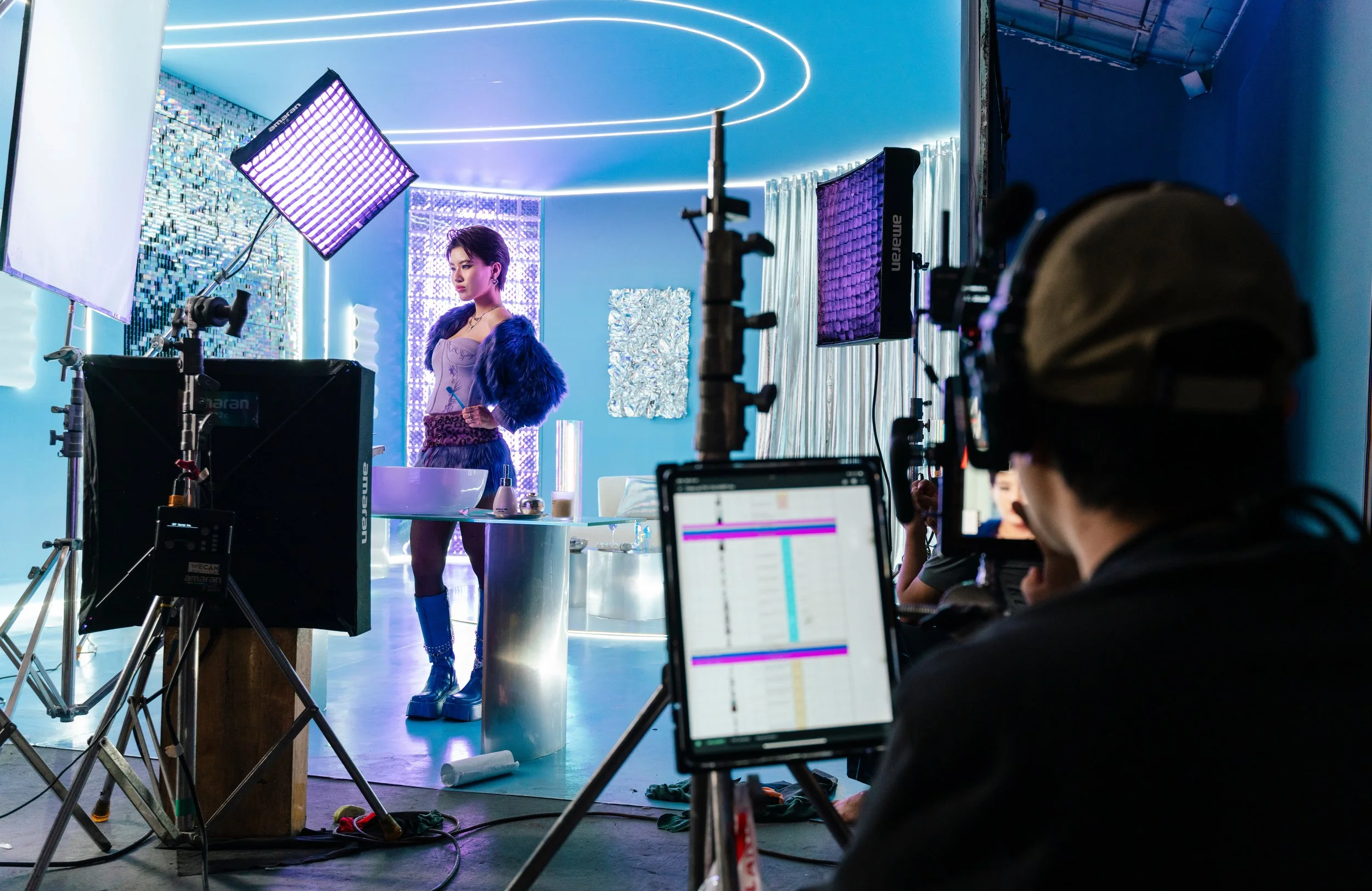 A woman posing in a colorful, modern set with professional lighting and filming equipment, preparing for a shoot or music video.