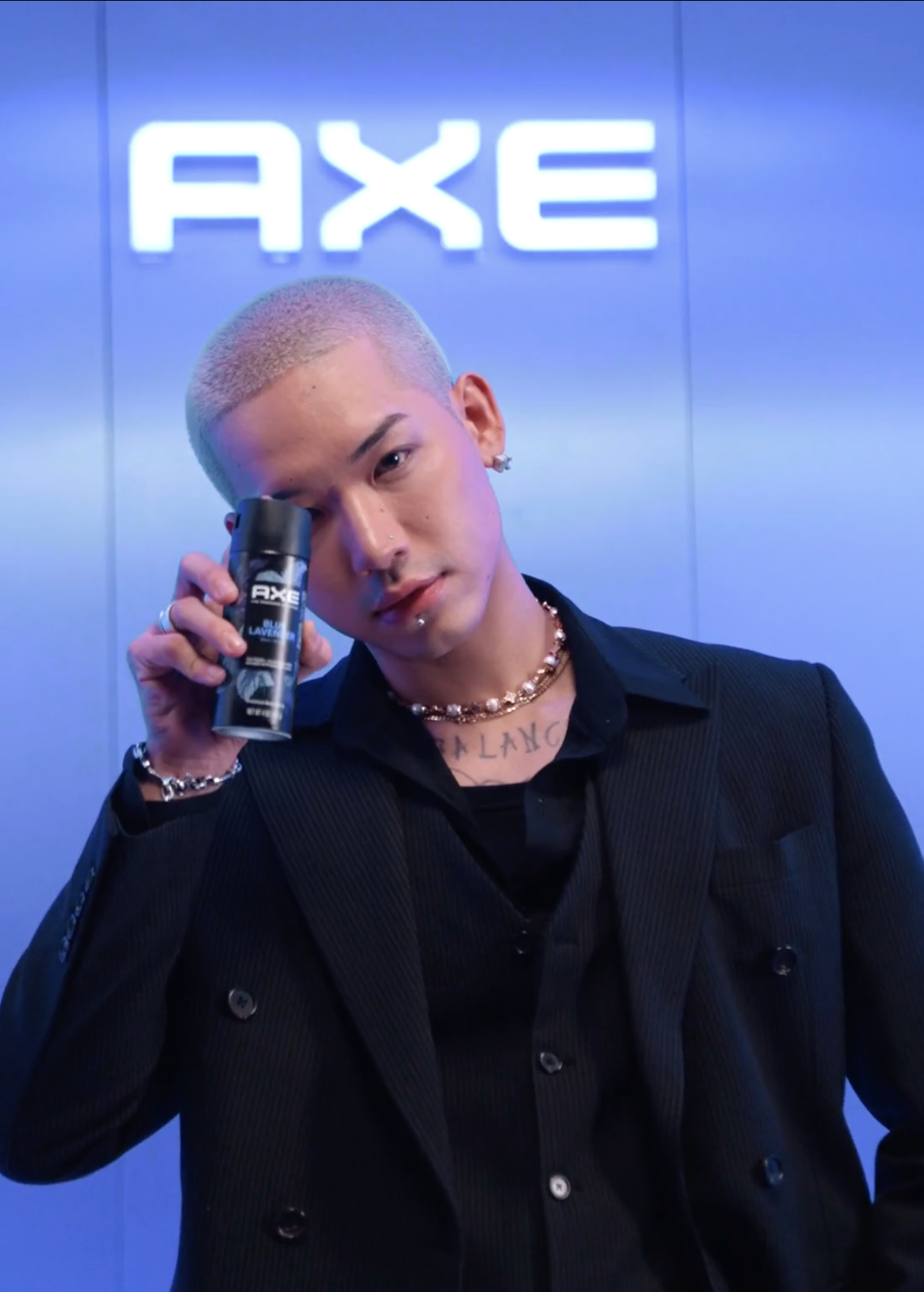 A young man with a buzz cut hairstyle holding a can of AXE body spray, standing in front of an AXE sign on a metallic wall.