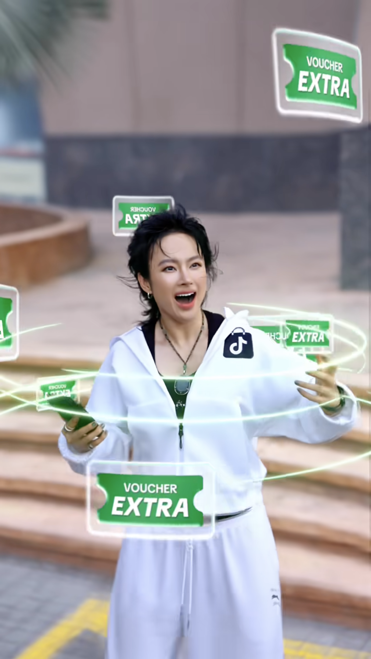 A young woman with short black hair, wearing a white tracksuit and jewelry, is holding a smartphone and looking surprised as green holographic vouchers labeled 'VOUCHER EXTRA' float around her.