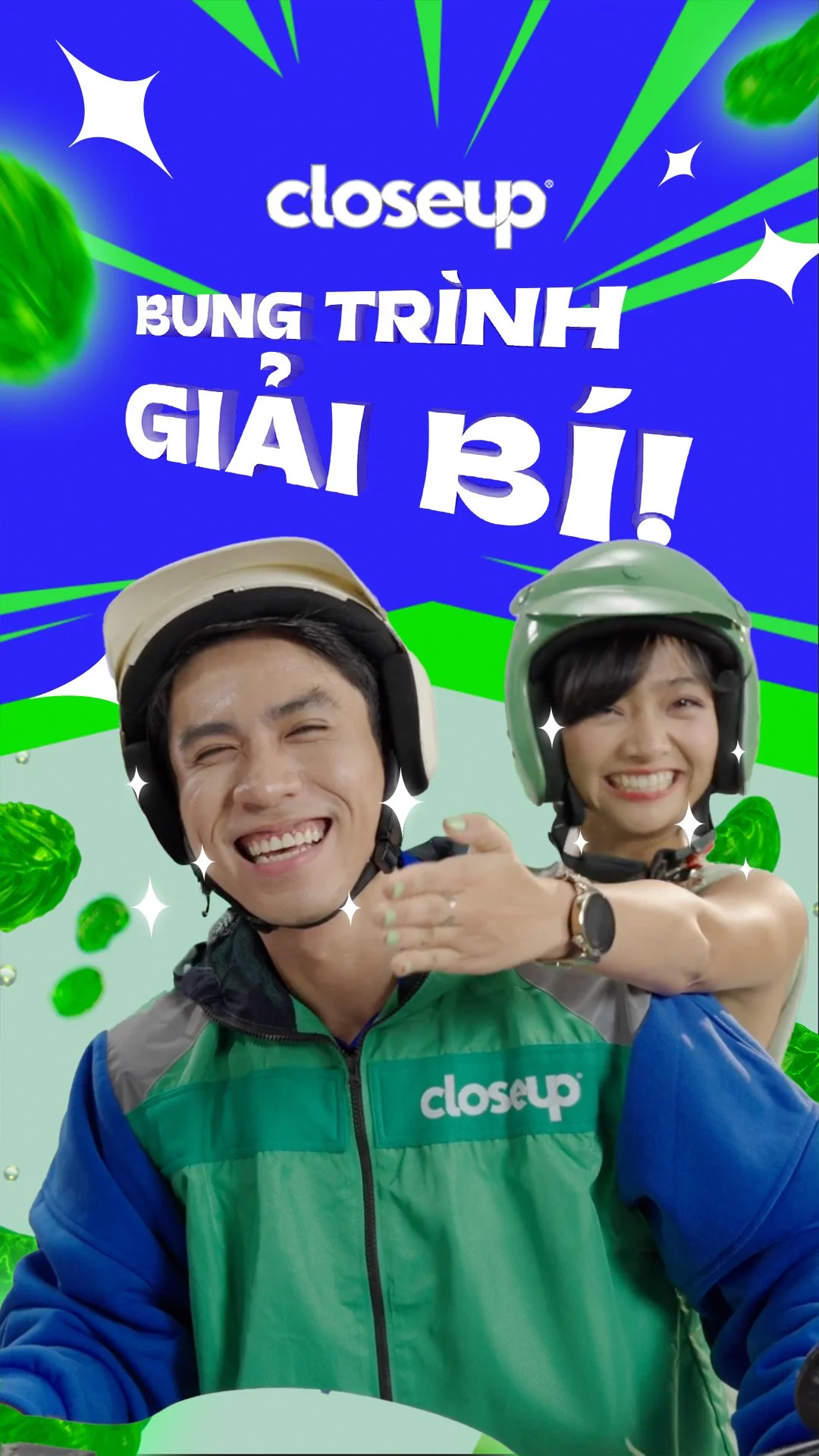 Two people wearing helmets and colorful jackets, smiling brightly. The background is vibrant with green and blue colors, featuring text in Vietnamese and playful graphics. The scene appears lively and cheerful, likely promoting a product or service.