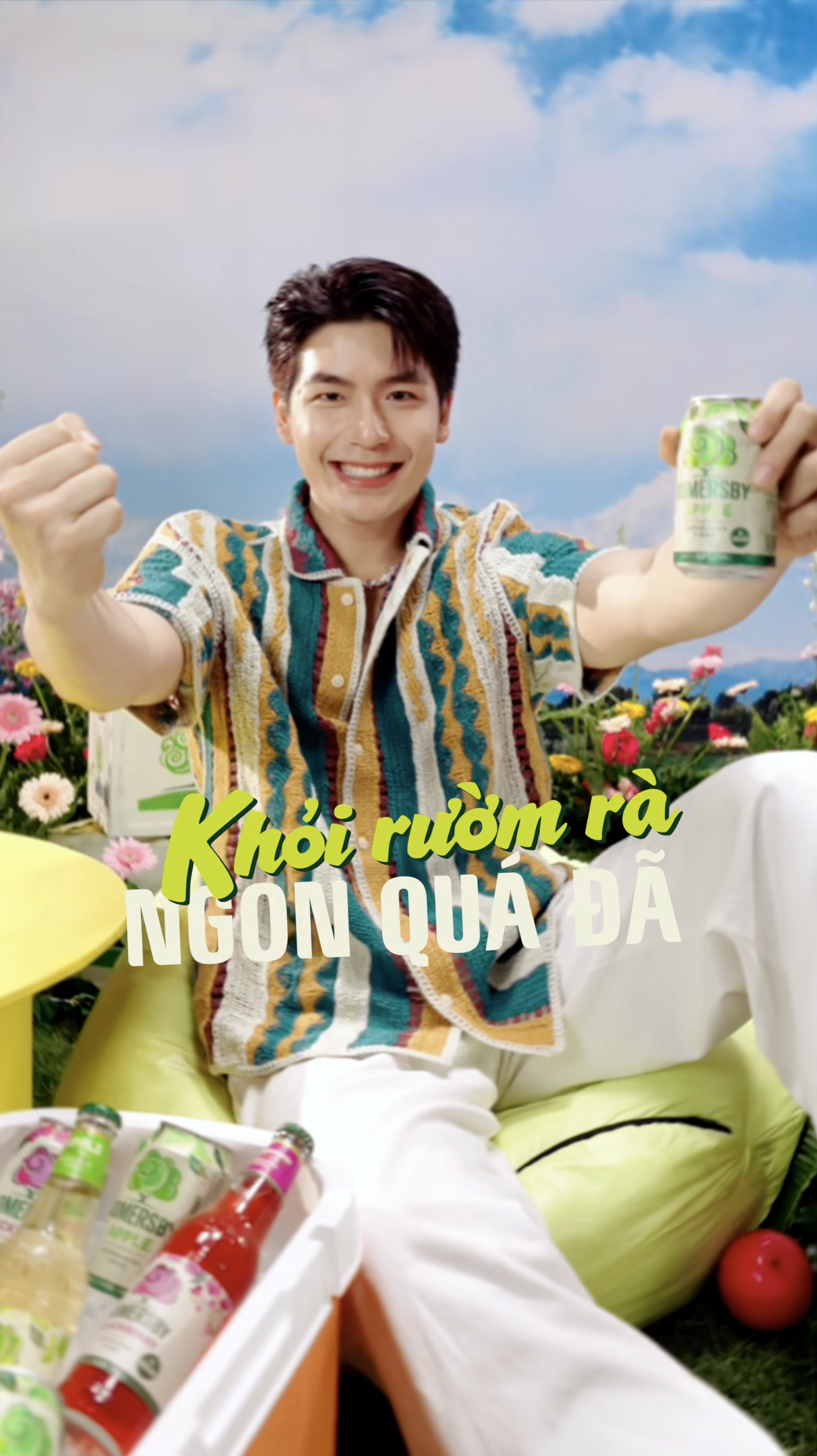 A young man in a colorful shirt smiling and holding a drink, sitting outdoors with flowers, a field, and blue sky in the background.