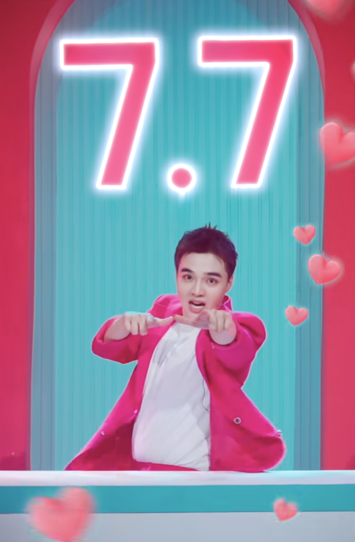 A young man in a pink blazer pointing towards the camera with a surprised expression, in front of a colorful background with a large neon sign reading '7.7' and floating pink hearts.