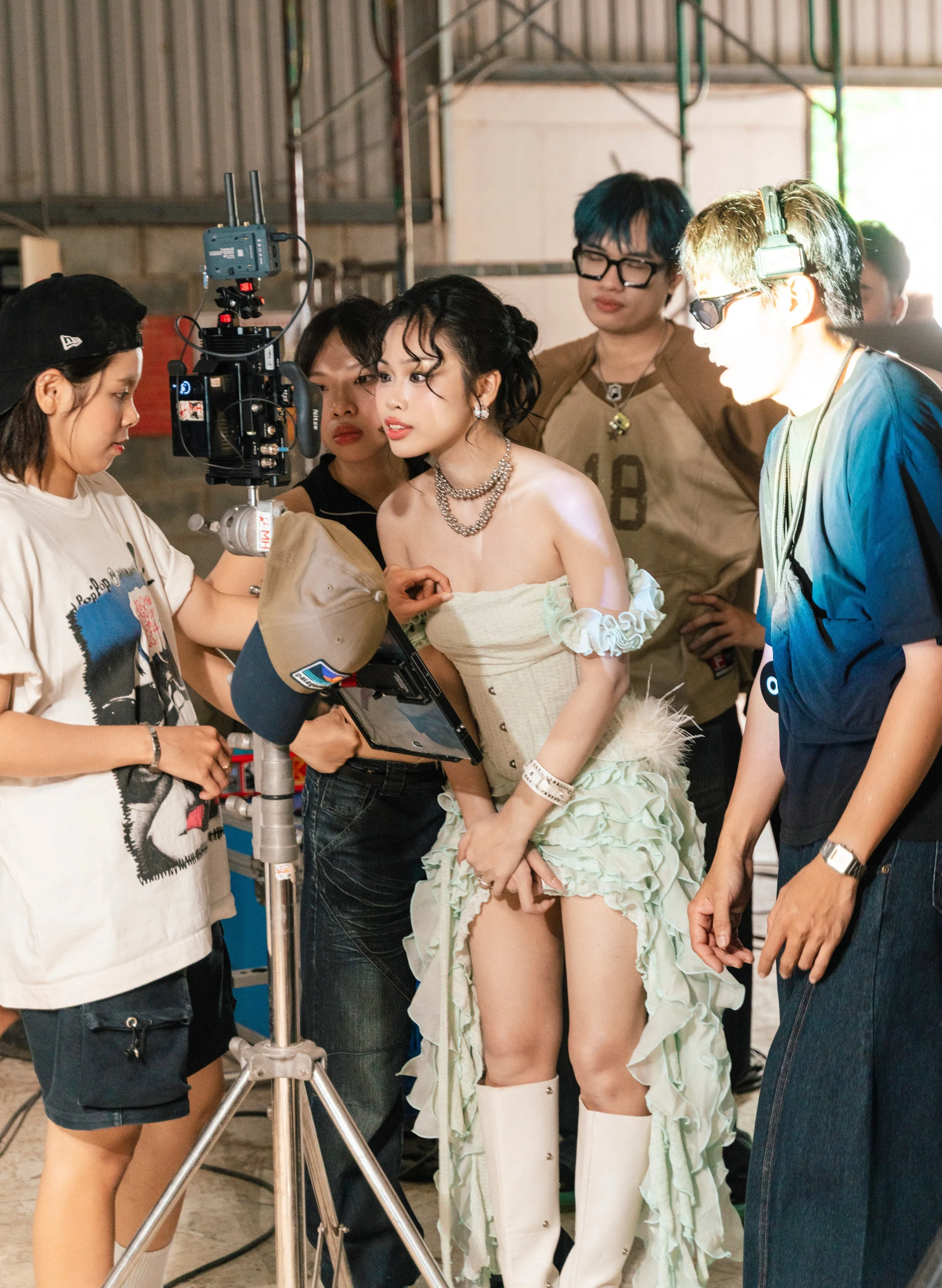 A woman on a film set, dressed in a unique outfit with accessories, stands in front of a camera. Several crew members are gathered around her, adjusting equipment and talking, with a factory-like or industrial background.
