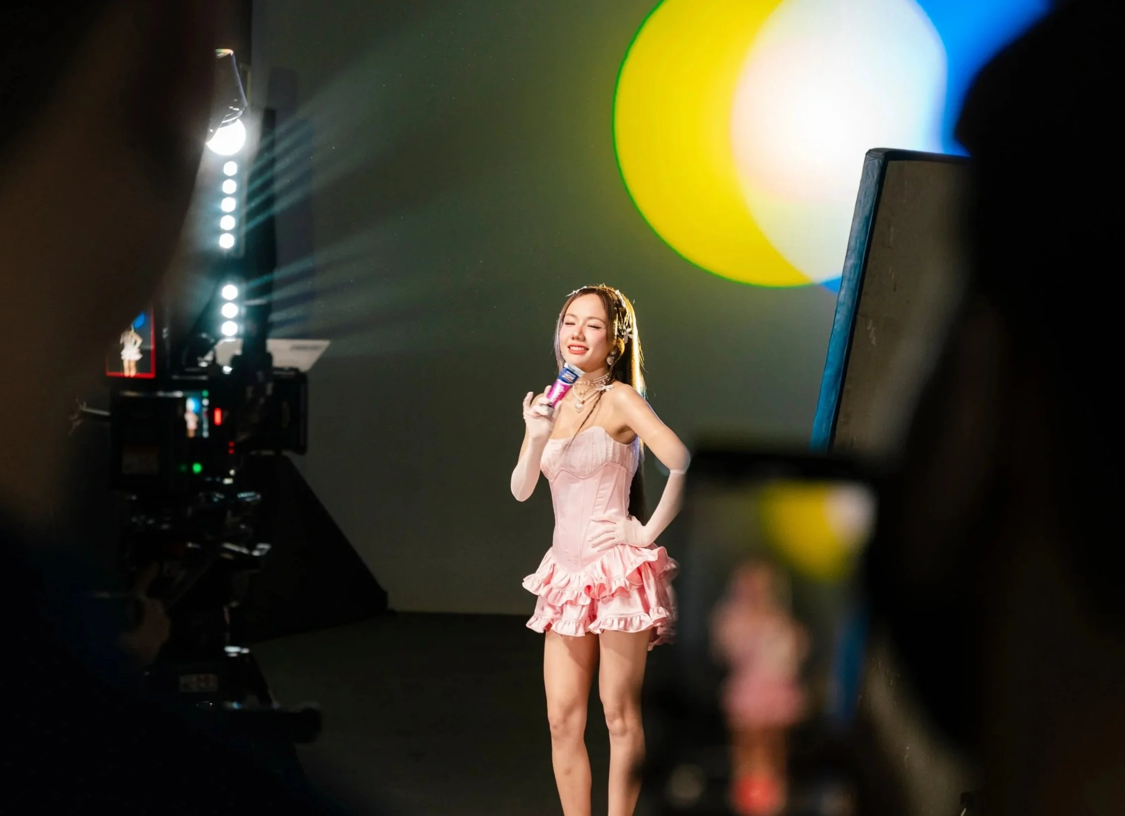 Young woman in pink ruffled dress singing into a microphone during a photo or video shoot, with camera equipment and studio lighting around her.