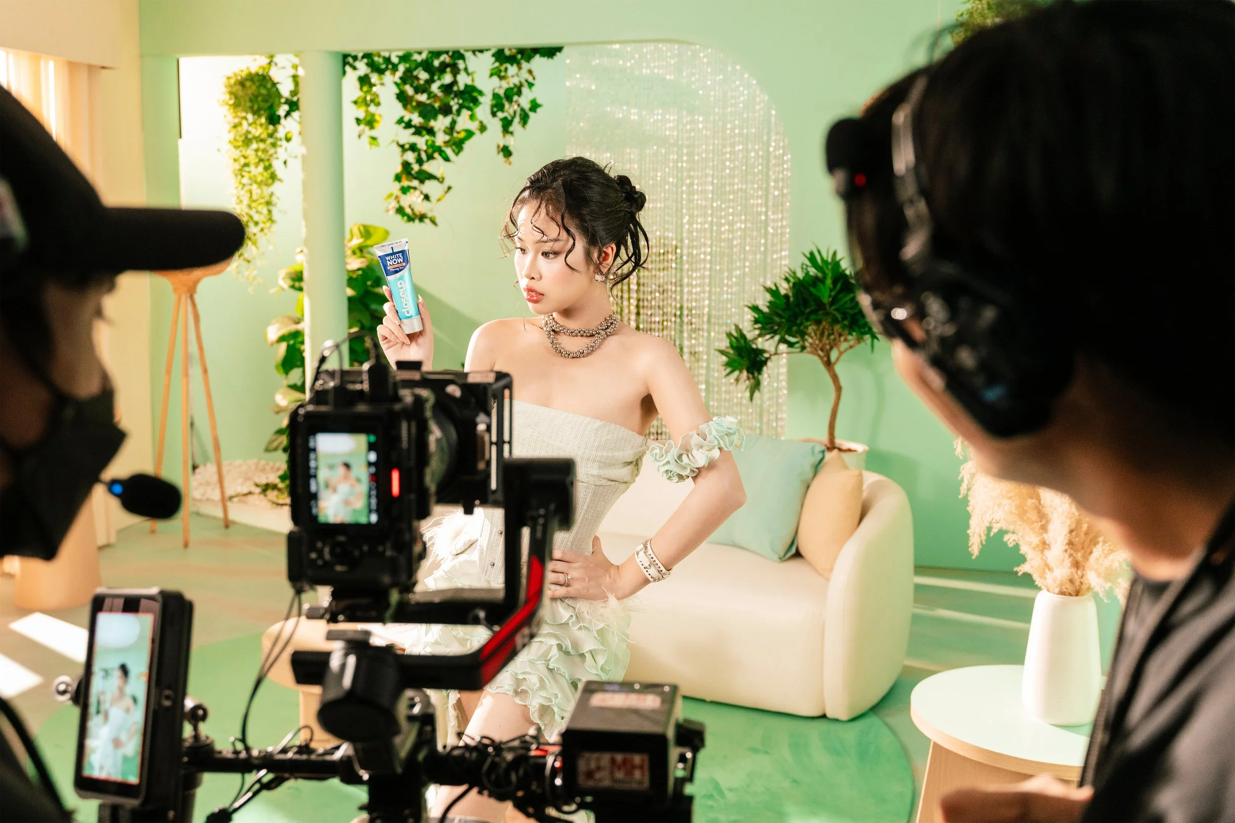 A woman in a white dress and jewelry posing for a photo or video shoot in a decorated green studio, holding a product in her hand, with two crew members operating cameras.