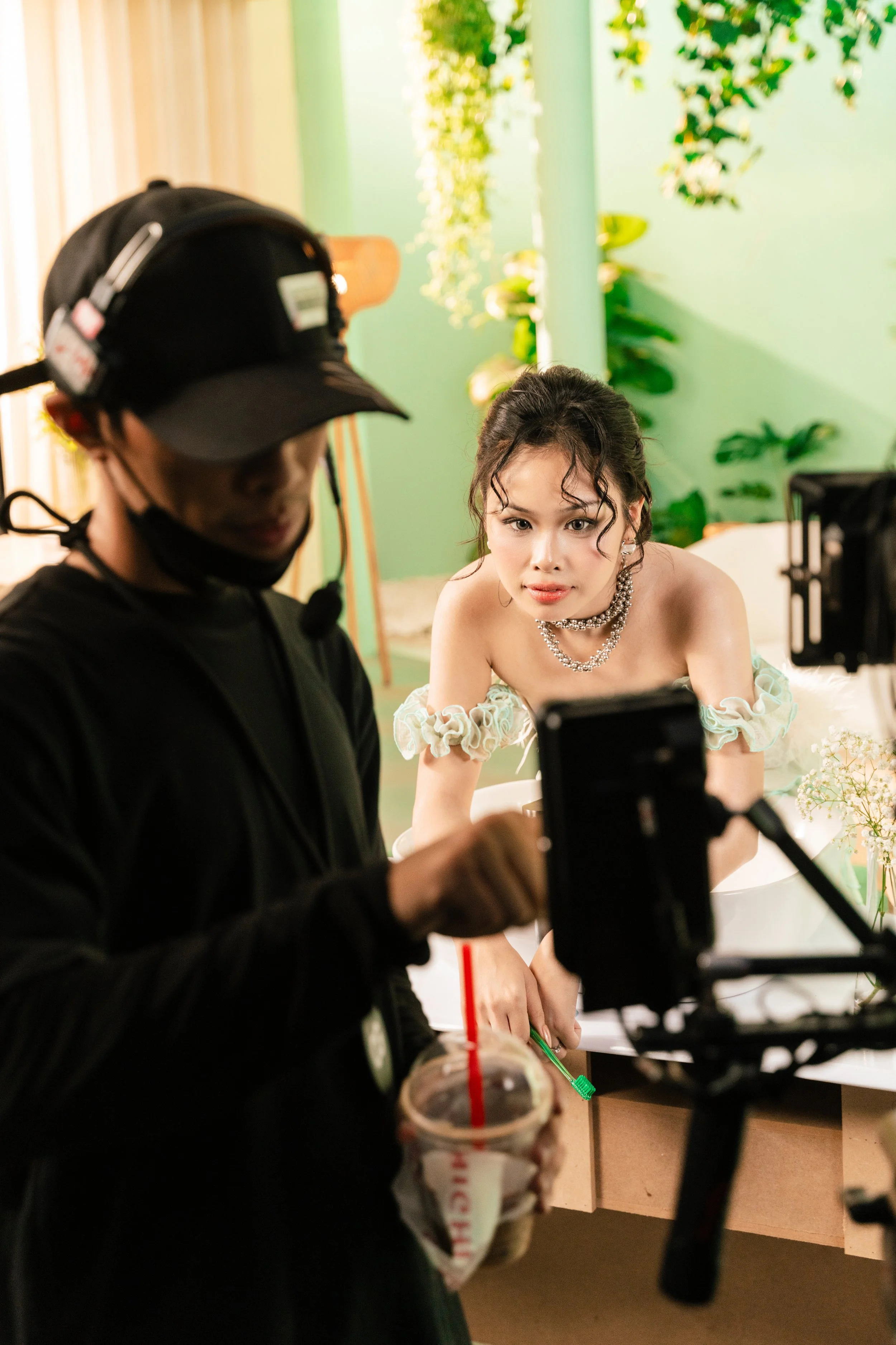 A woman with curly hair, wearing a floral off-shoulder dress and layered necklaces, looks at a camera on a filming set. A person in dark clothing holding a drink is in the foreground.