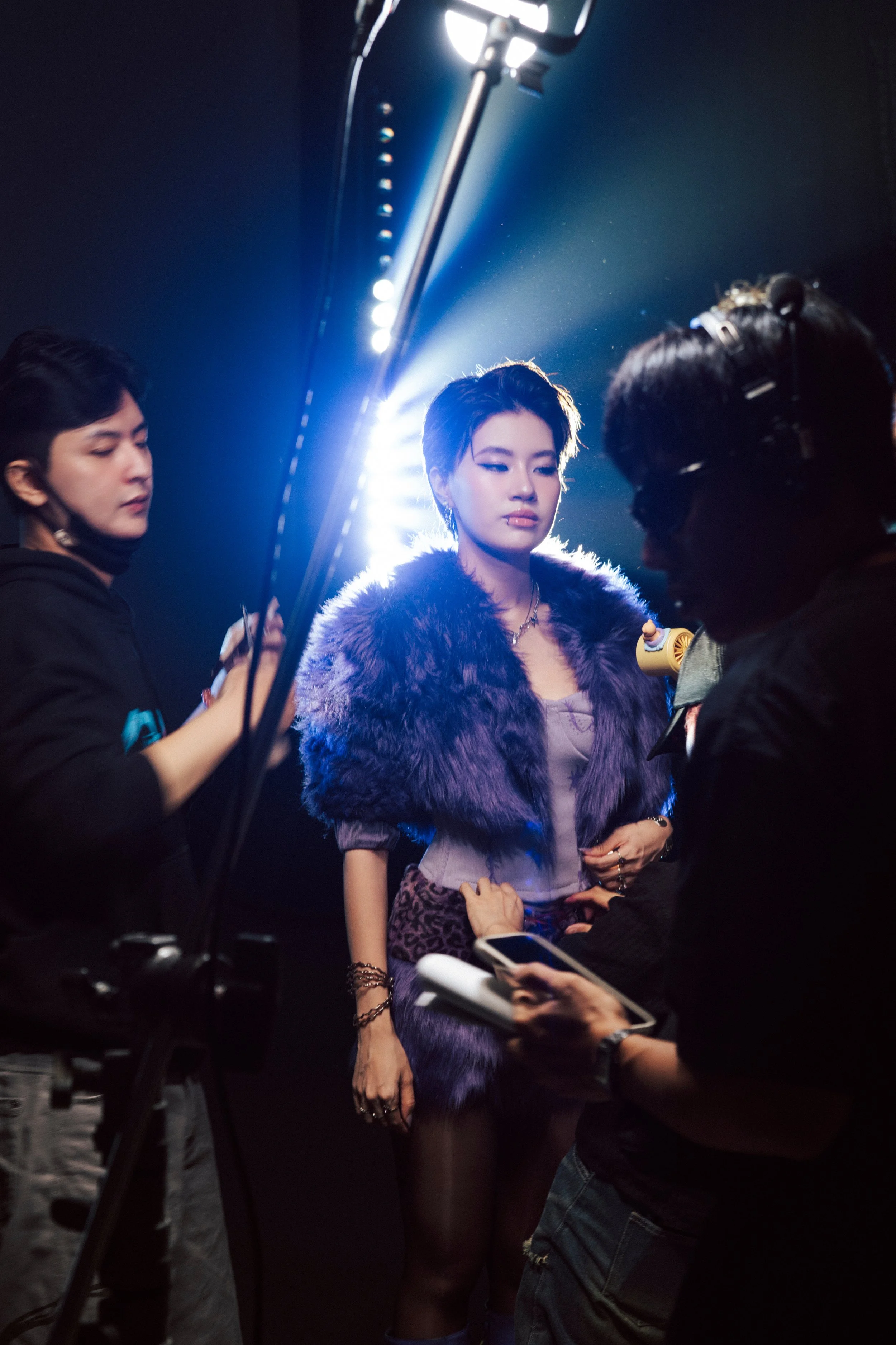 A woman in a purple fur stole and leopard print skirt being assisted by crew members on a film set with bright studio lights.