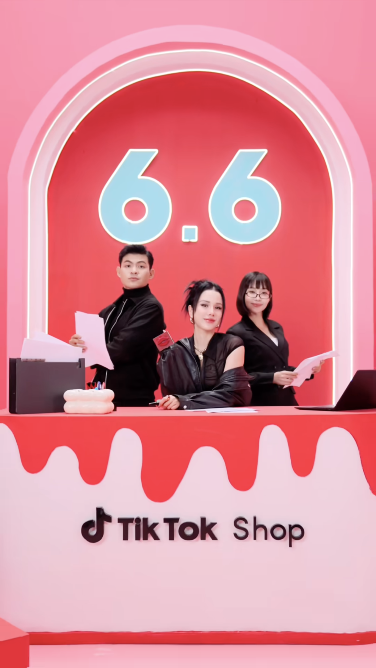 Three people standing behind a TikTok shop counter with a large red and pink background displaying the number 6.6. The person in the middle is a woman with black hair and a black leather jacket, seated at the table, while the two others, a man and a woman, are standing behind her, holding papers. The shop has a pink and red theme with a large TikTok logo and the words 'TikTok Shop' on the front.