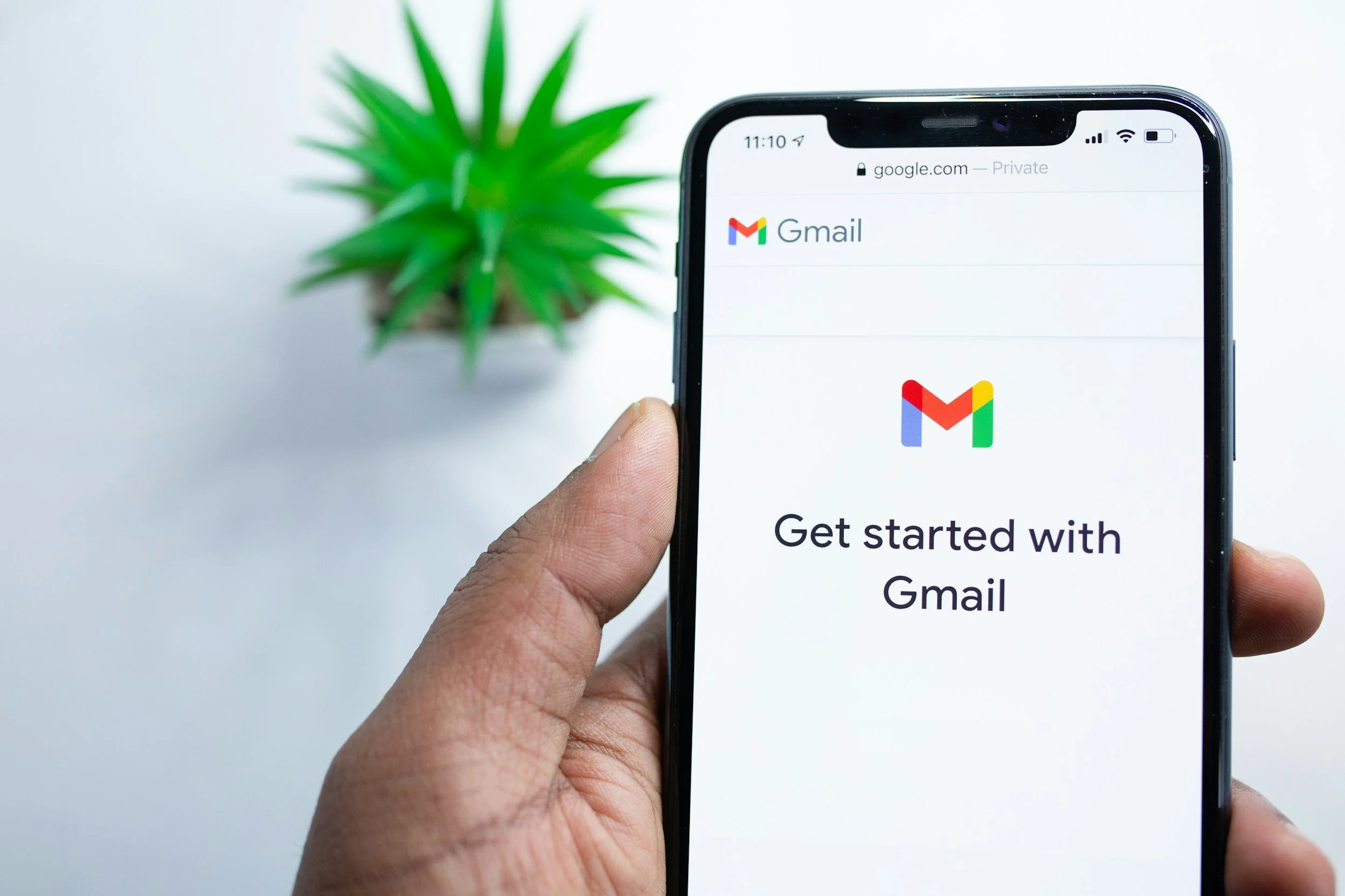 Person holding a smartphone displaying the Gmail app with the message 'Get started with Gmail' on the screen, with a potted green plant in the background.