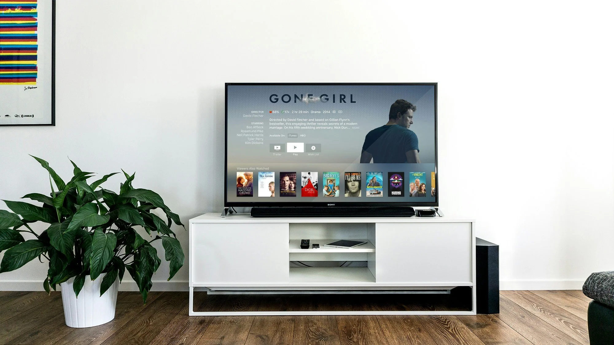 Living room with a Sony TV on a white stand, a large green potted plant, and a partially visible gray sofa. The TV screen displays a show called "Gone Girl" and a row of other movie thumbnails at the bottom.