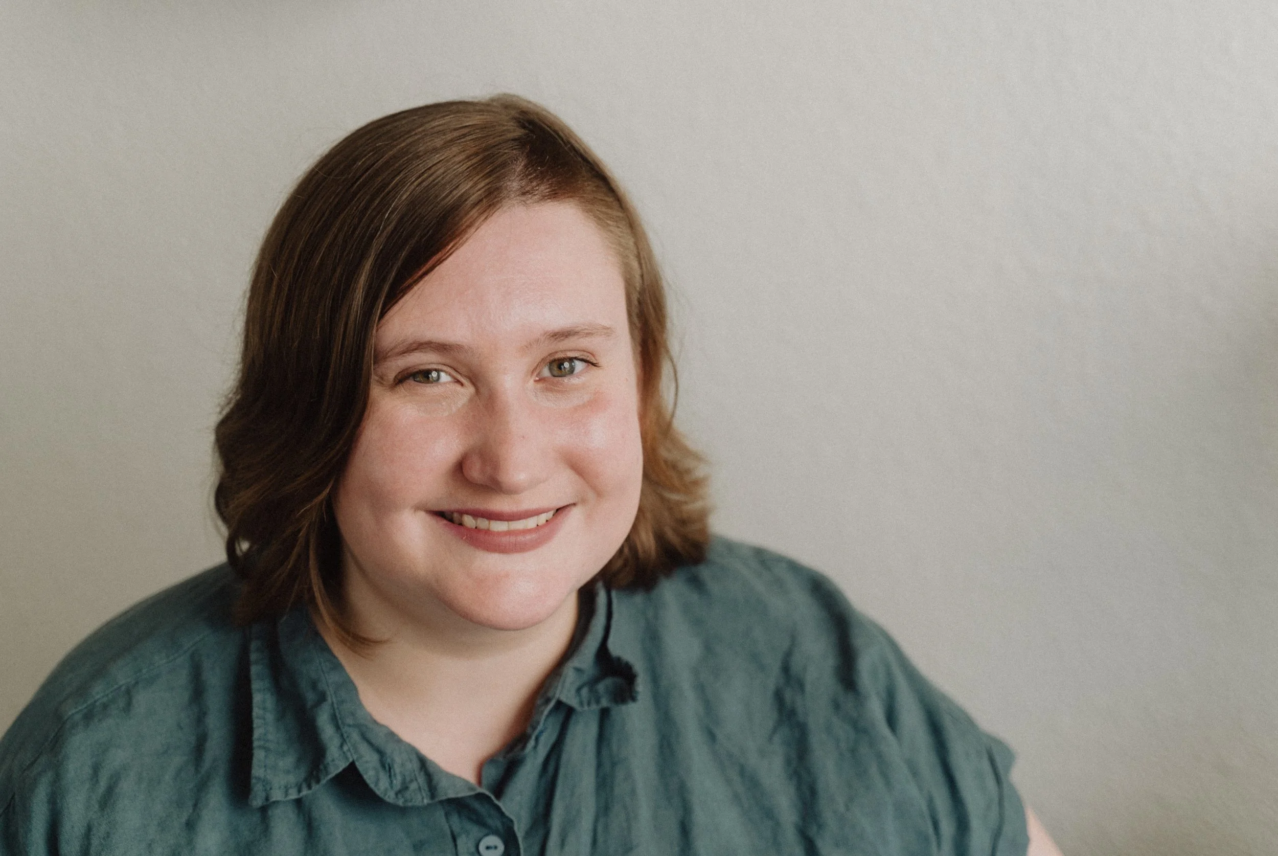Portrait of a smiling woman with short brown hair, wearing a dark green shirt, against a plain wall.