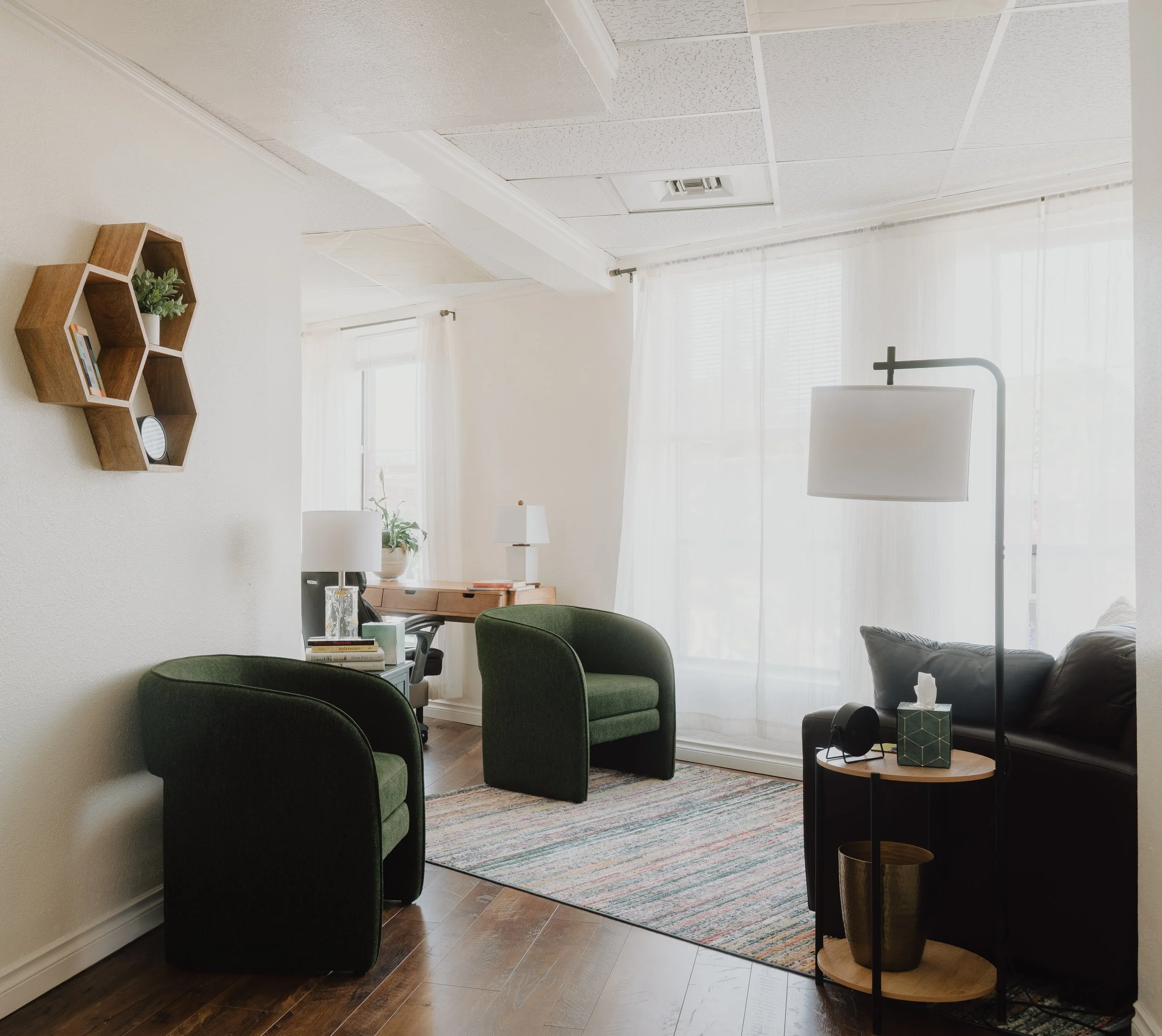 Picture of the office with two single chairs to the left, a couch to the right, and a desk in the far left corner. There are large windows with natural light