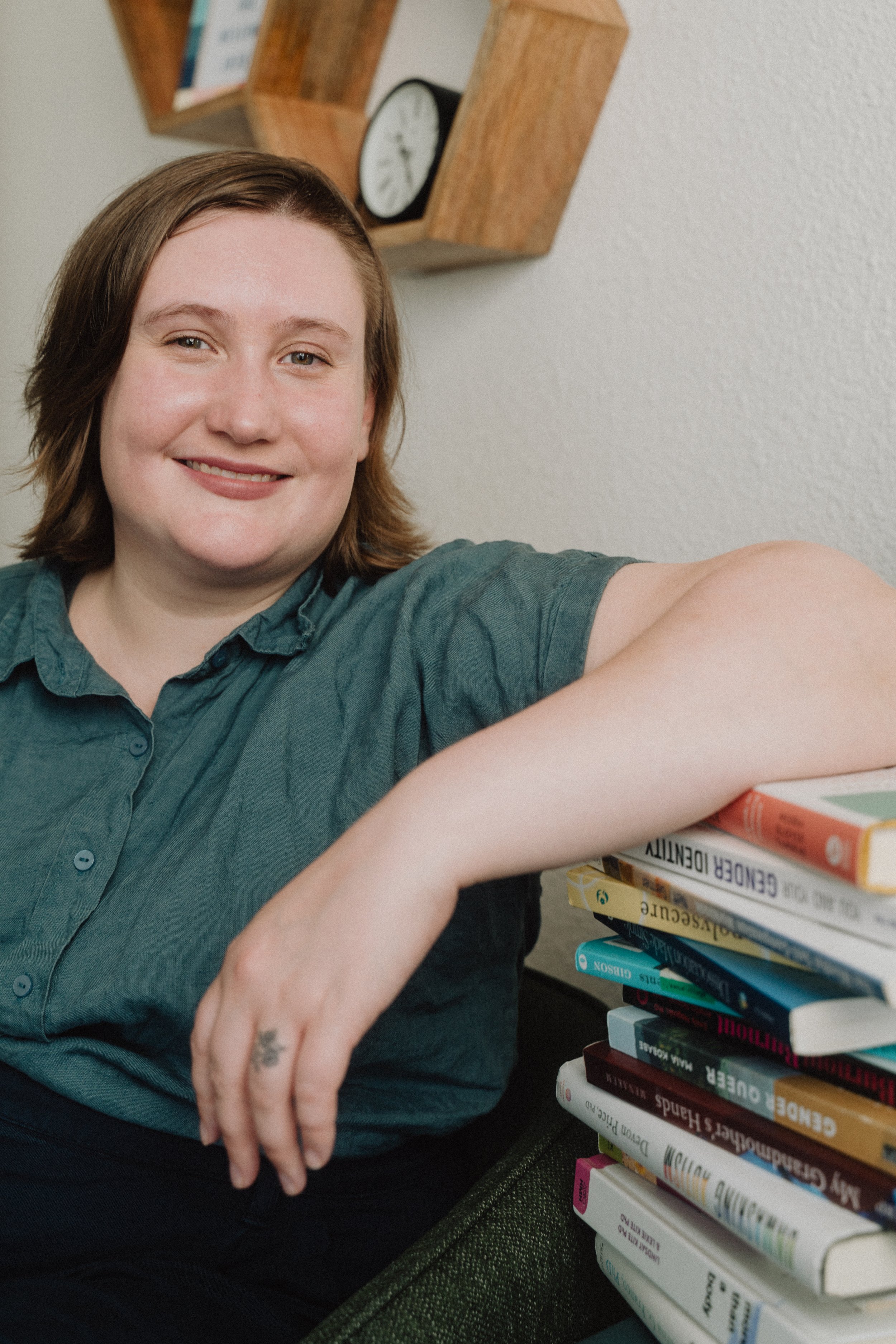 Young woman with short brown hair, wearing a green button-up shirt, smiling and sitting next to a stack of books on a desk.