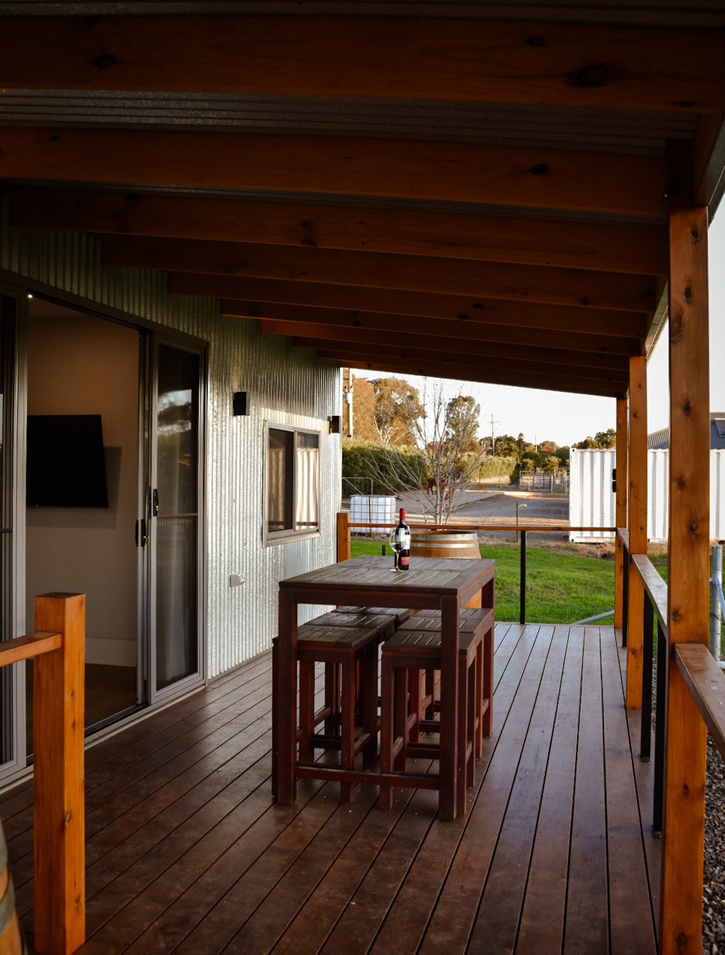 Wooden porch with a table, a bottle of wine, and two glasses, overlooking a grassy yard with trees in the background.