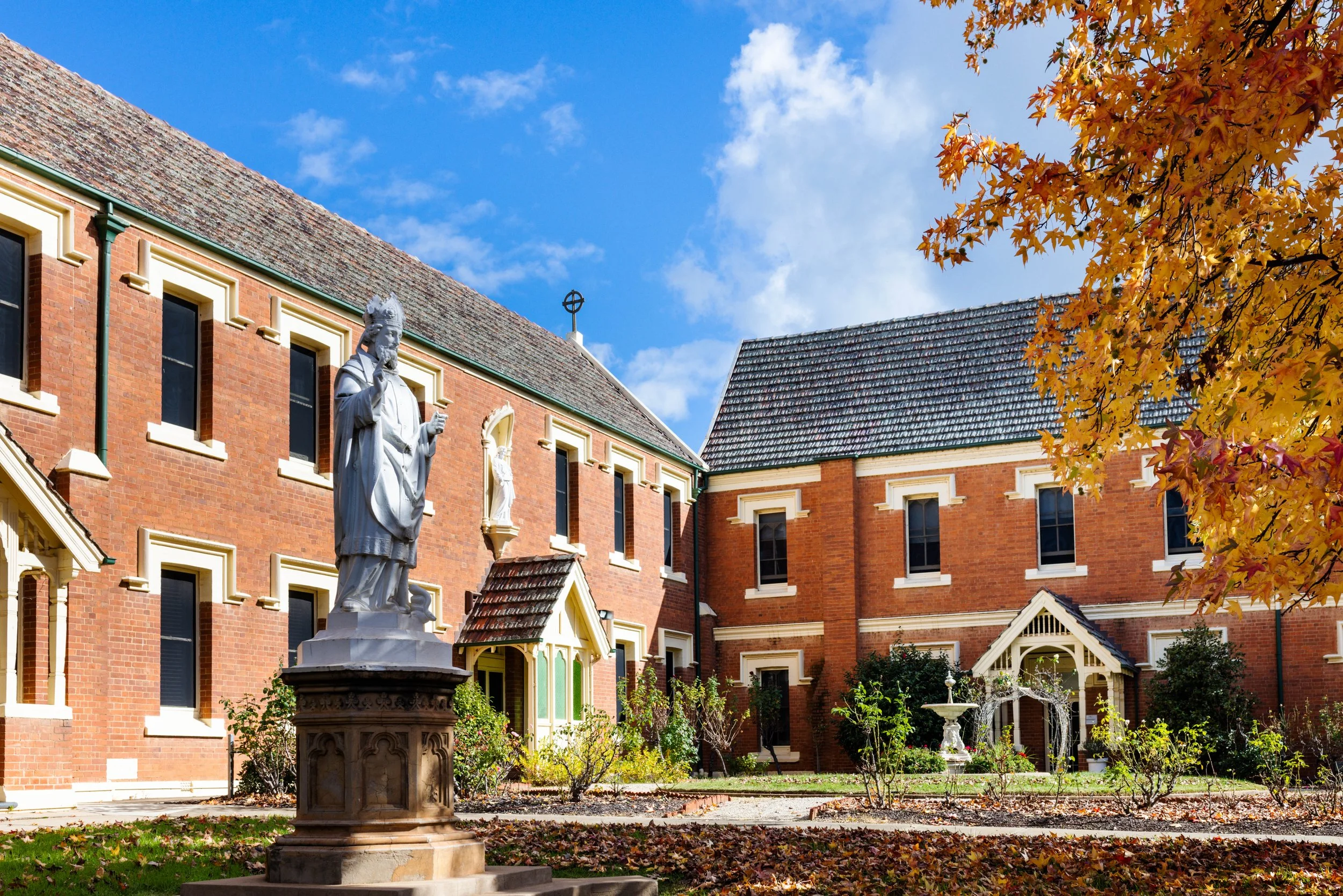 A courtyard with a statue of a religious figure on a pedestal, surrounded by red brick buildings with white trim, a tree with orange leaves, and a blue sky with clouds.