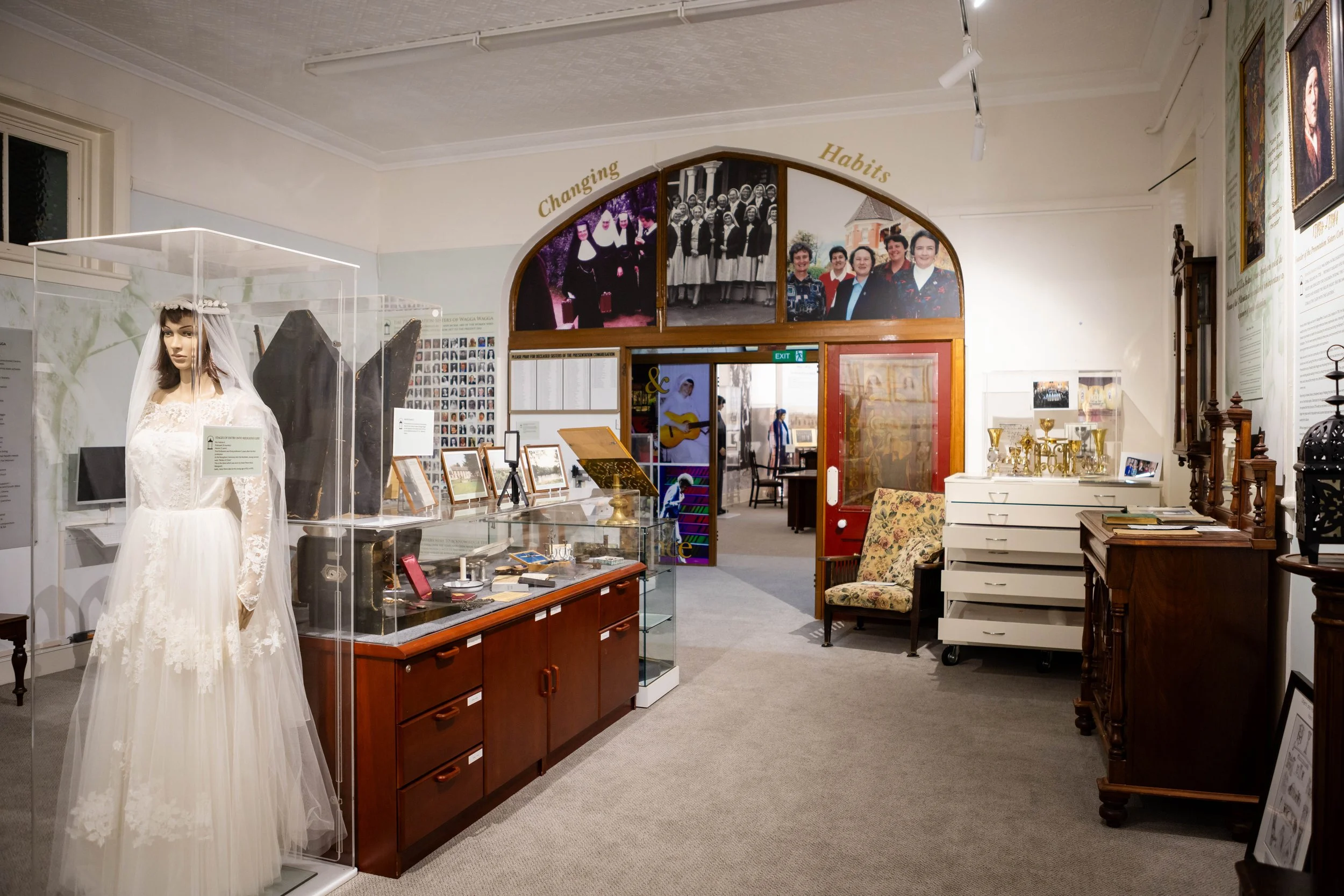 A museum exhibit featuring a mannequin dressed in a vintage wedding gown with a veil, displayed in a glass case. Behind the gown, there are photographs and informational displays on the wall, including a black-and-white group photo, a color photo of women posing outdoors, and portraits of historical figures.