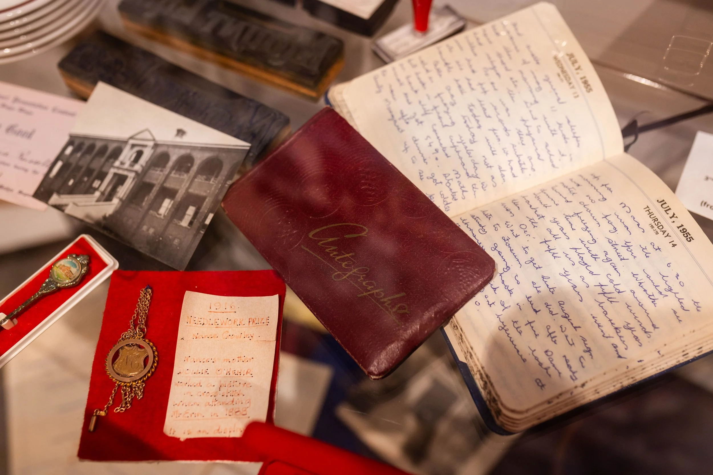 A collection of vintage items on display, including handwritten letters, a red book, and jewelry with a medal or badge, along with a black and white photograph of a building.