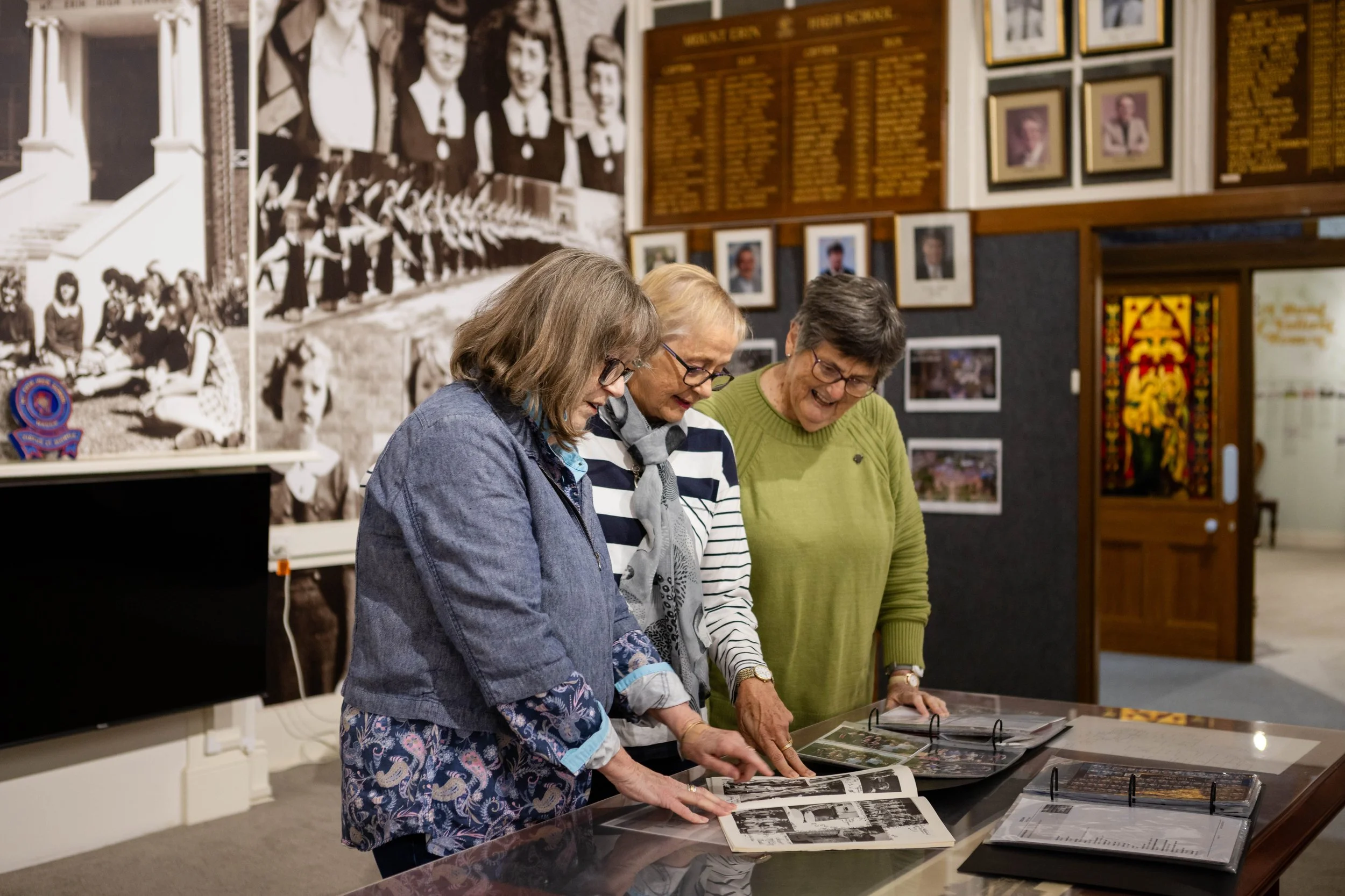 Three elderly women with glasses looking at photo albums in a museum or historical exhibit with framed pictures and historical memorabilia on the walls.