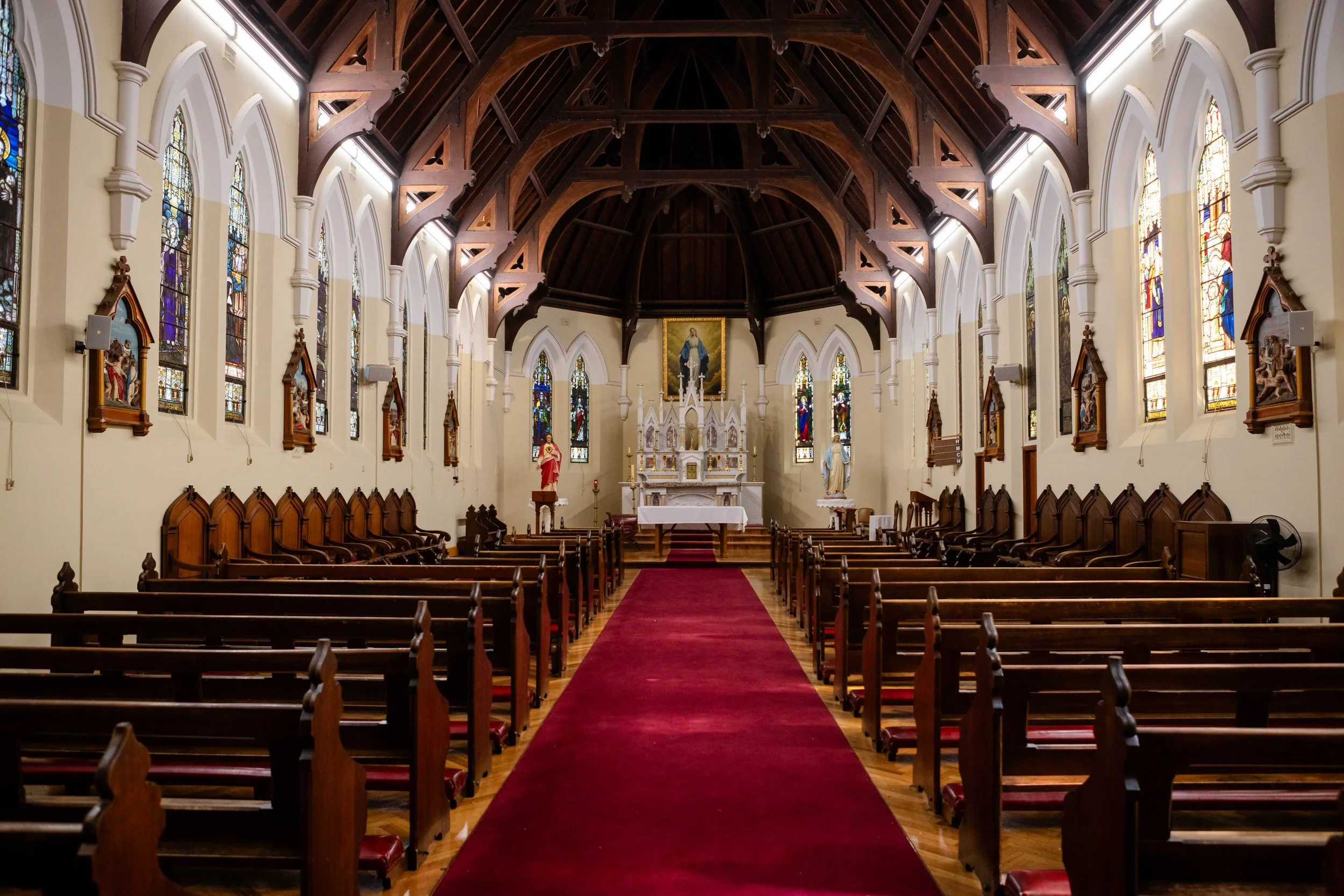 Interior of a church with wooden pews, stained glass windows, and religious statues along the walls. Central aisle covered with red carpet leading to the altar.
