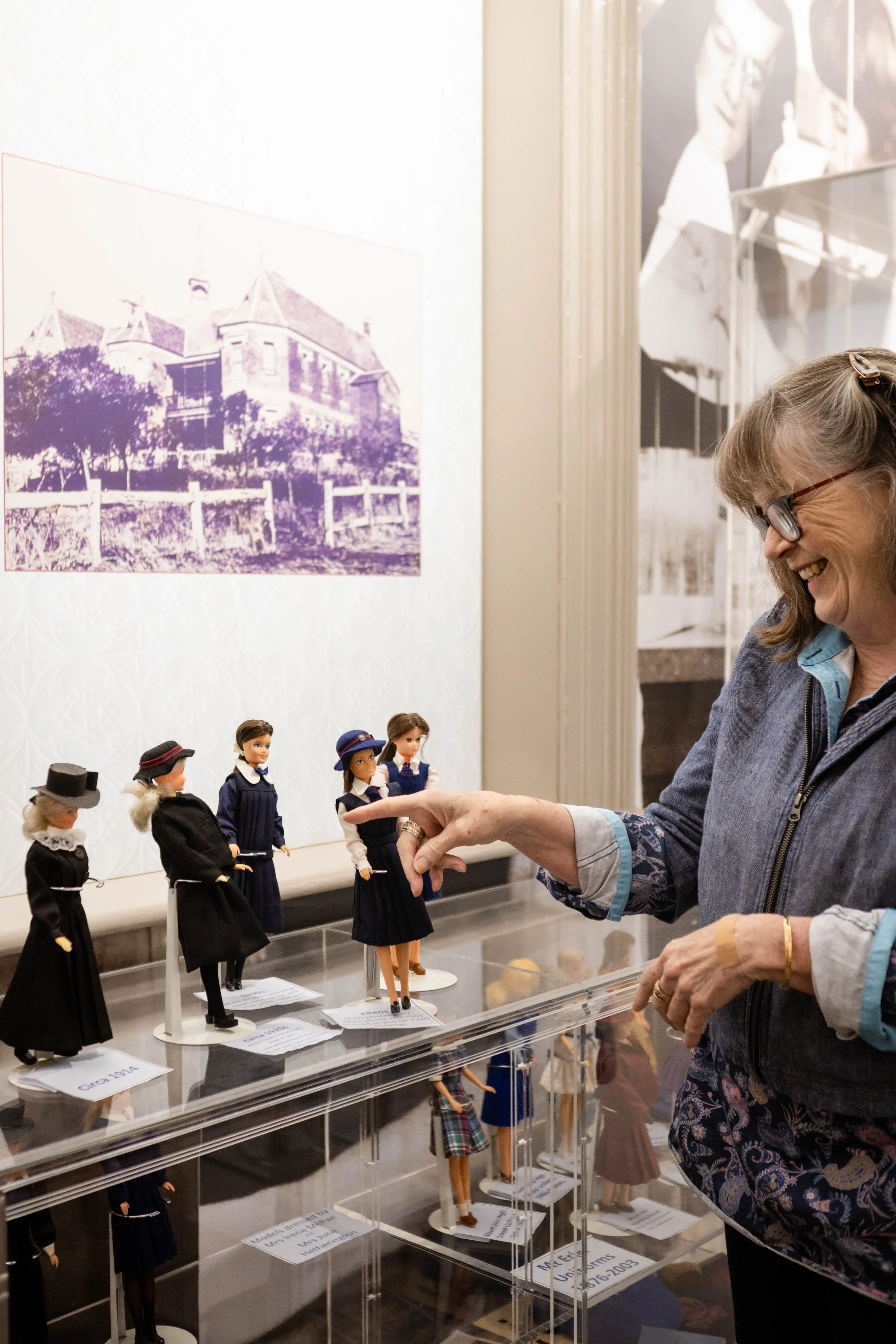 A woman smiling and wearing glasses looks at a display of vintage doll clothing and mannequins dressed in historical costumes in a museum exhibit.