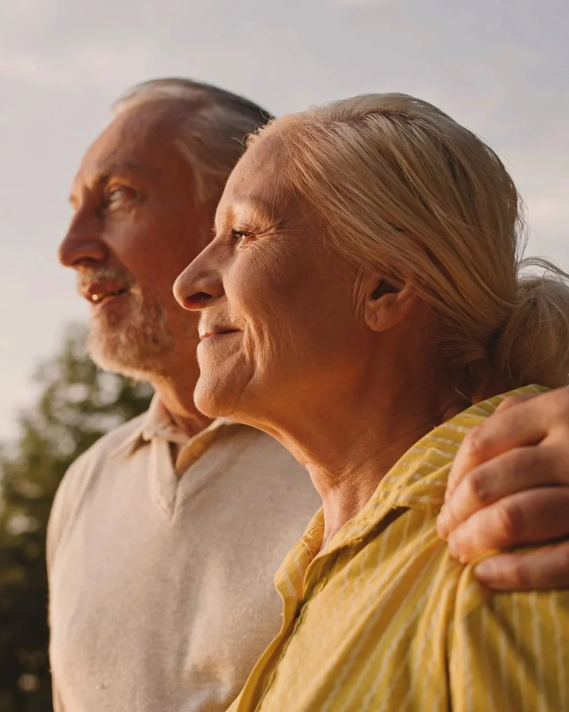 An elderly man and woman are standing outdoors, smiling and embracing each other while looking into the distance during sunset.