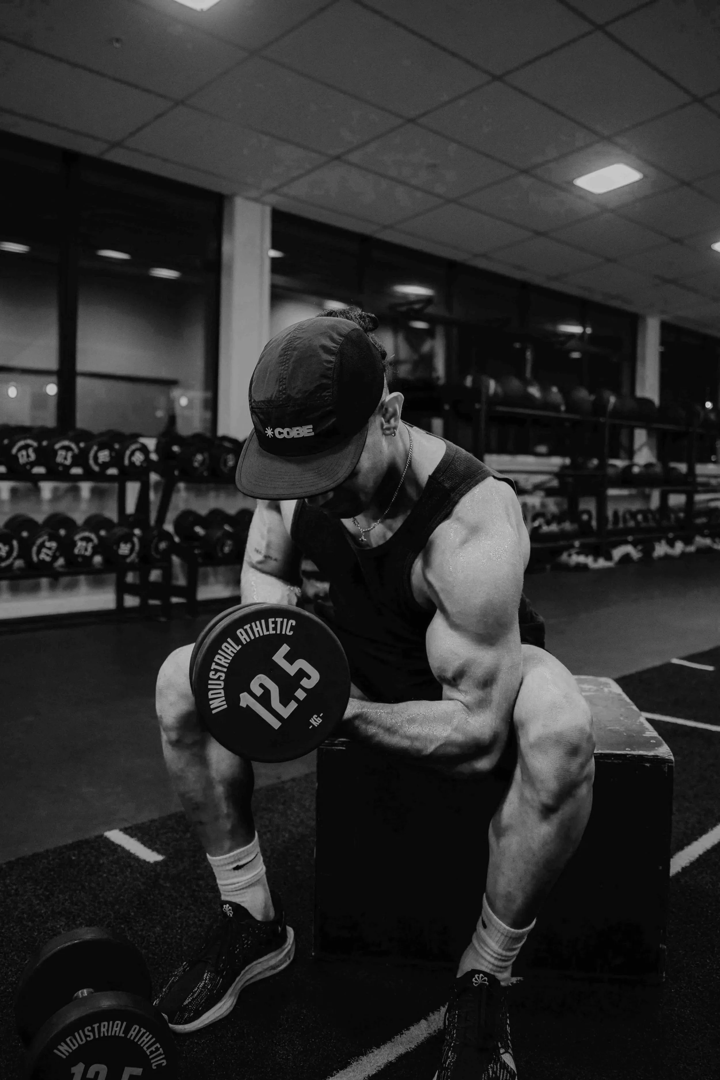 A person in a gym is bending over to lift a weight plate labeled '12.5' with the words 'INDUSTRIAL ATHLETIC' on it. The individual is wearing a cap, sleeveless shirt, and compression socks. In the background, there are shelves with multiple weights and a row of gym equipment.