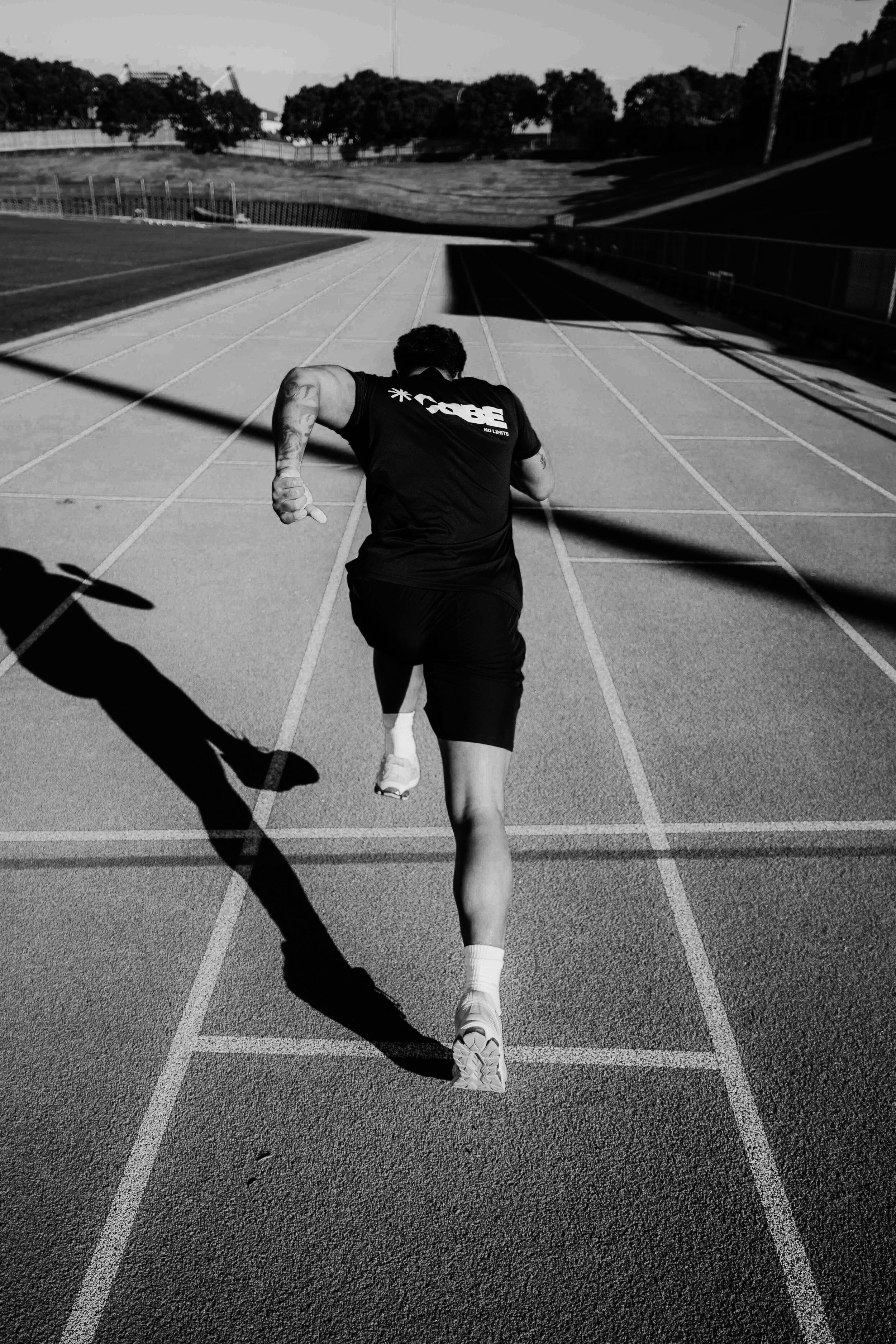 A man running on an outdoor track with a hill and trees in the background, seen from behind in black and white.