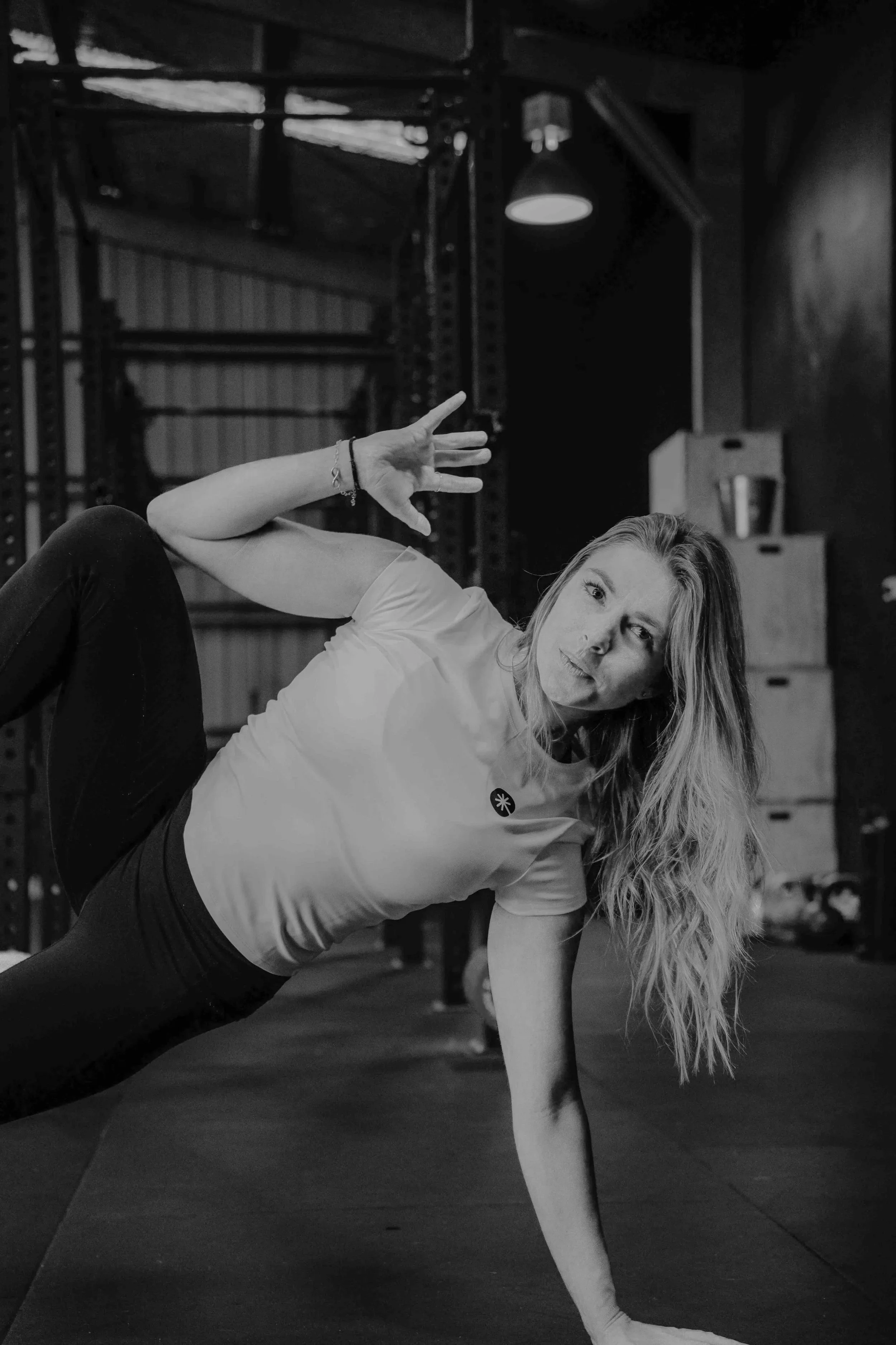 A woman with long wavy hair doing a side plank exercise in a gym, supporting herself with one arm and raising the other hand with fingers spread. The gym has metal racks and boxes in the background.