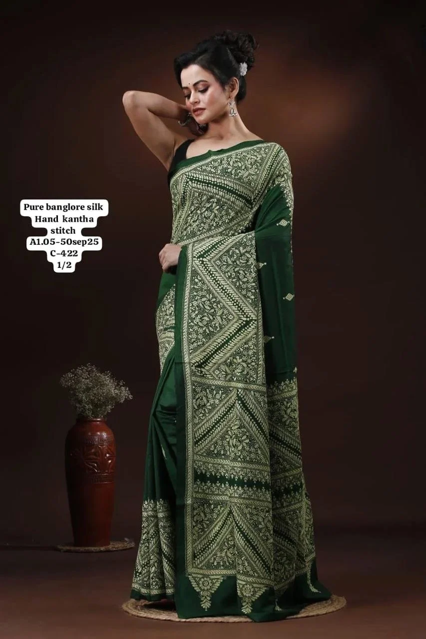 A woman posed in a traditional green and gold Indian saree made of pure Bangalore silk, featuring hand Kantha stitches. She stands with one hand behind her head, with dark hair styled up, and accessorized with earrings. A red vase with dried flowers is on the floor beside her against a dark background.