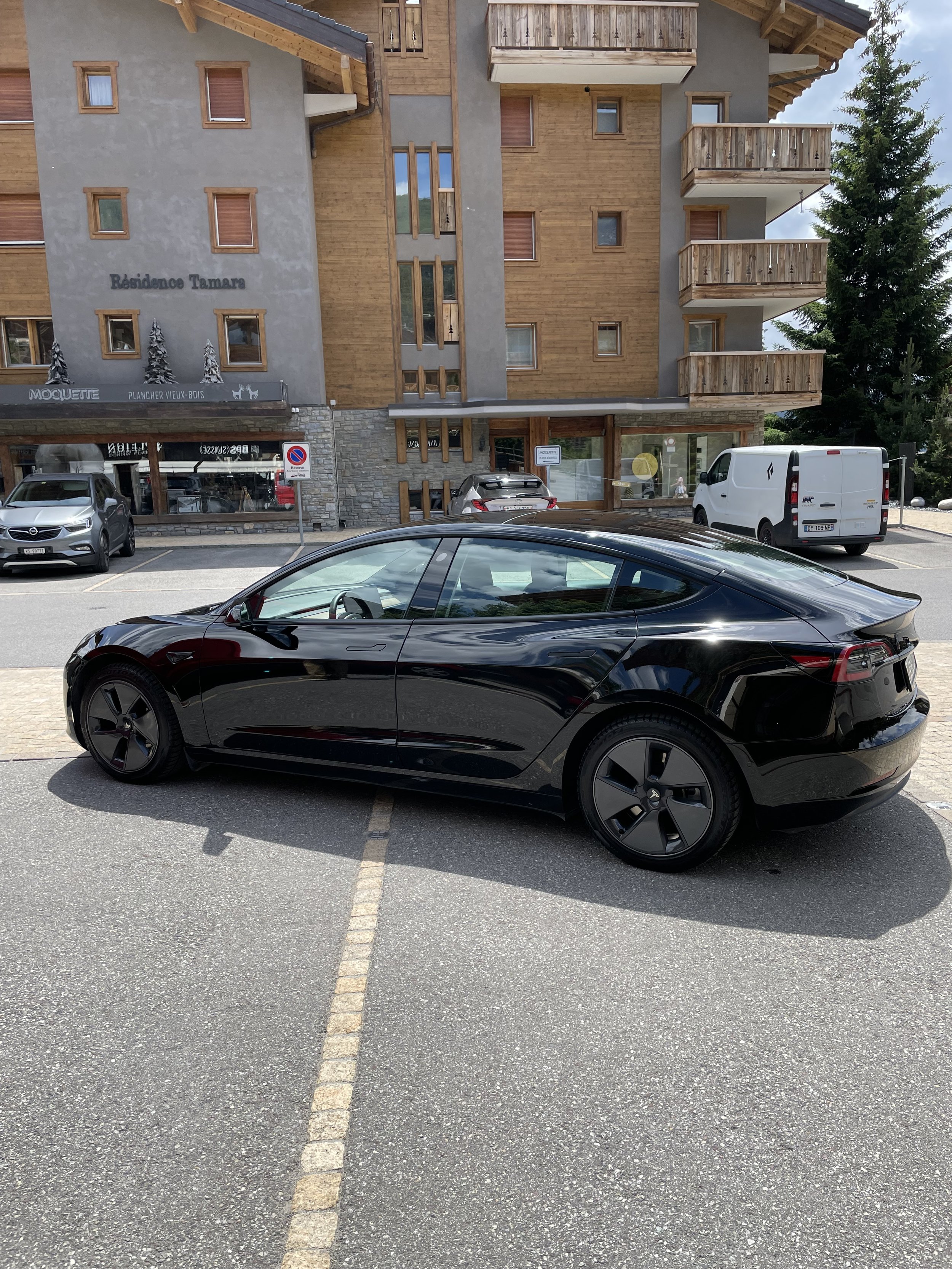 A black Tesla parked in a marked parking space in front of a multi-story building with wooden balconies and windows, with other vehicles and a tree in the background.