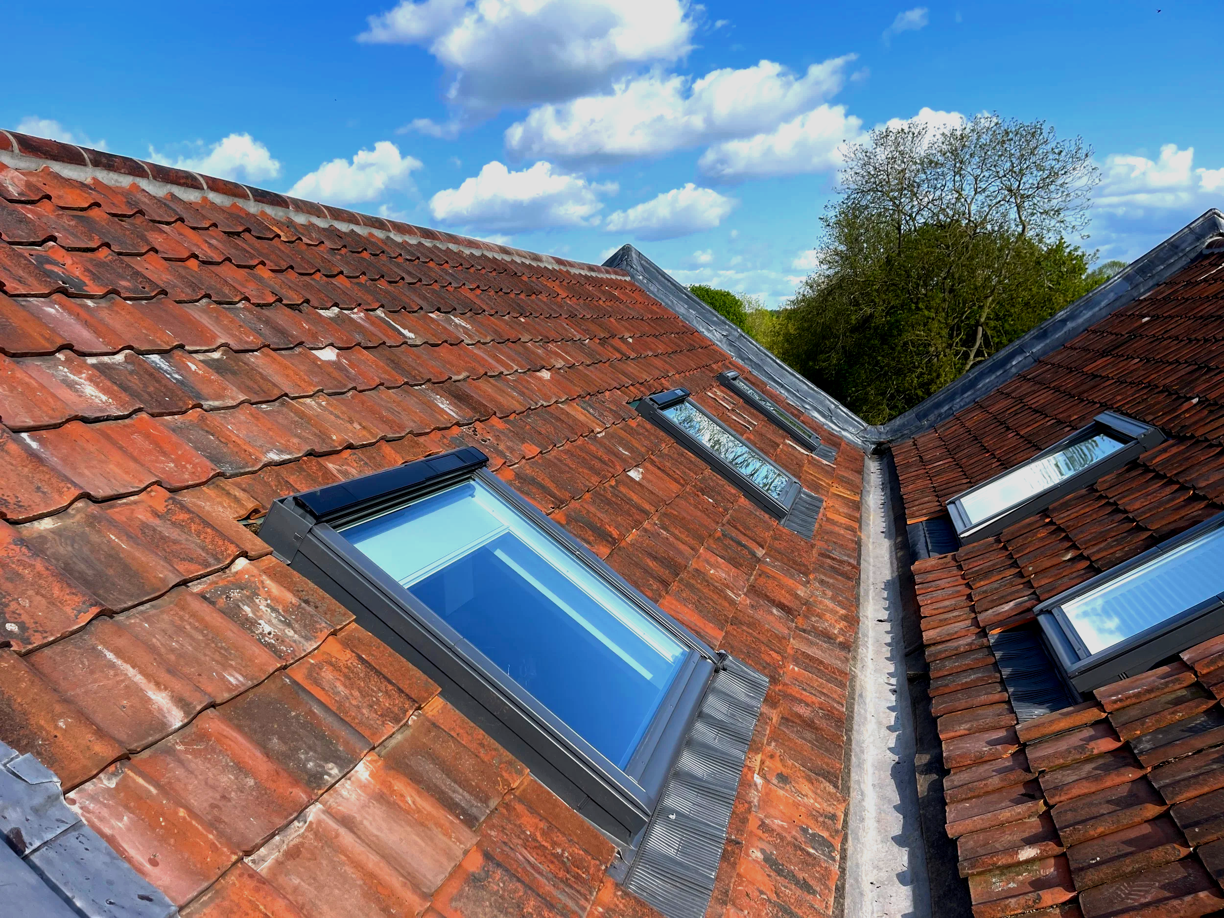View of a red-tiled roof with several skylights, and trees against a partly cloudy sky.