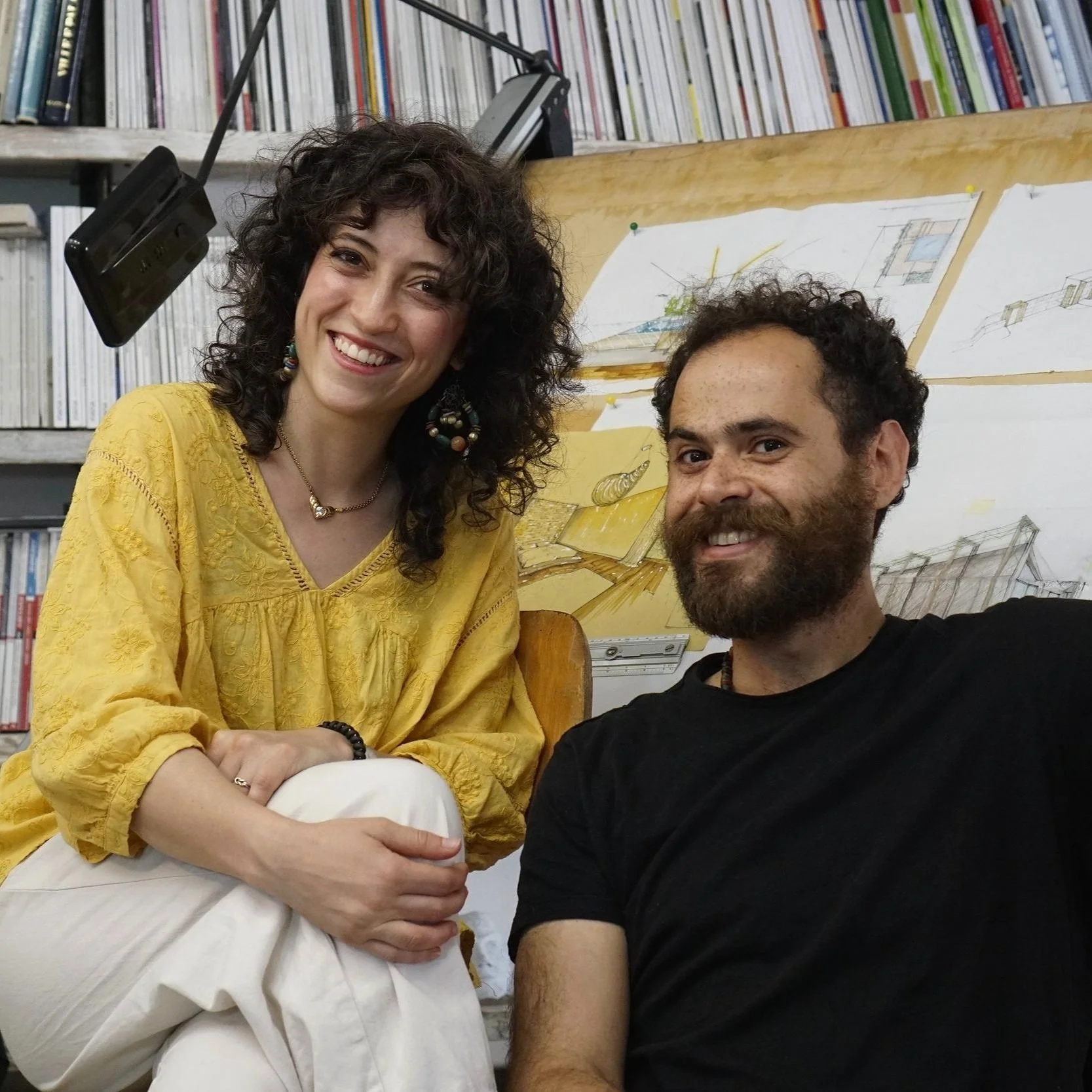 Una donna con capelli ricci e vestito giallo sorridente, seduta accanto a un uomo con barba e capelli ricci, entrambi sorridenti, con libri e disegni di architettura sullo sfondo.
