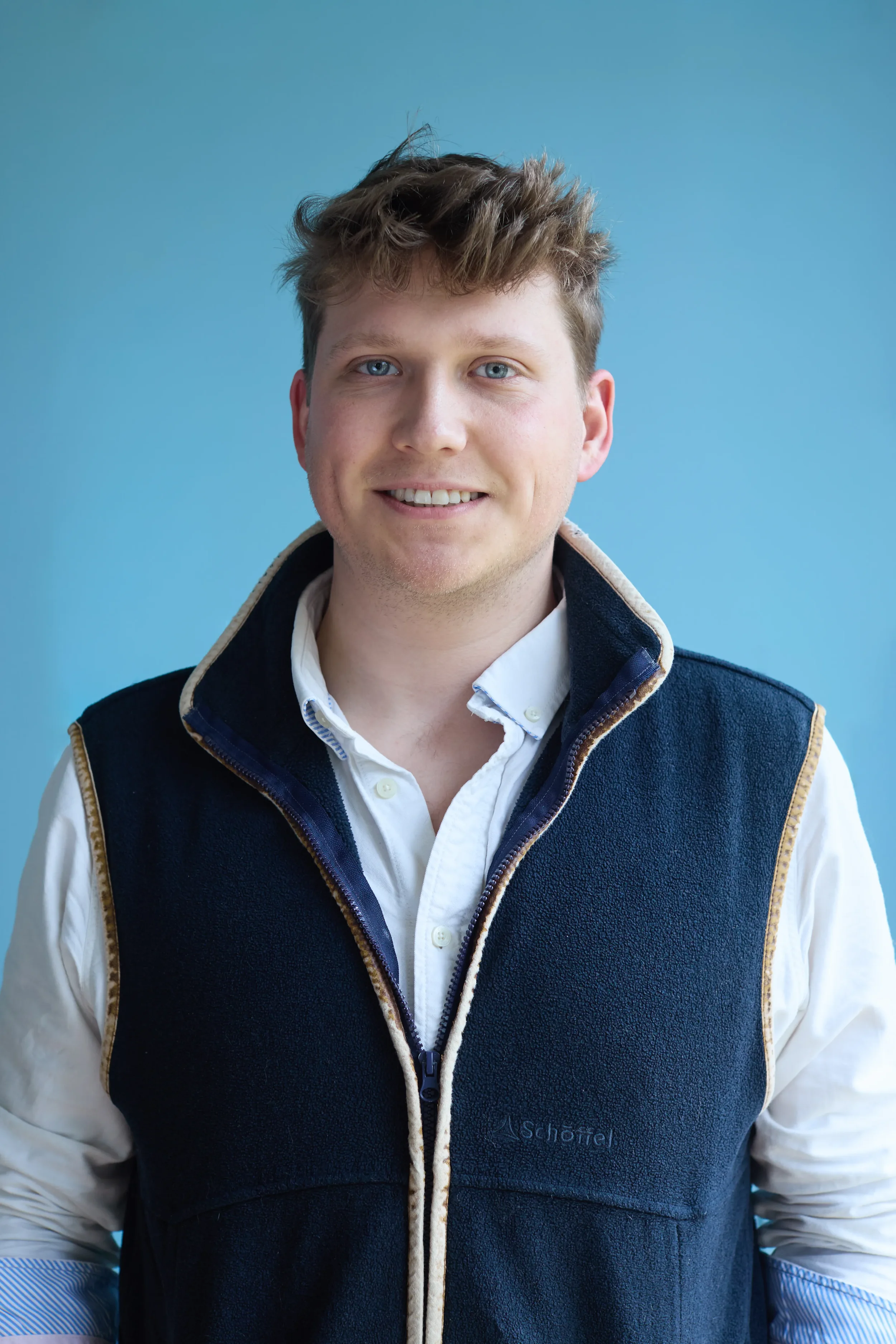 A young man with light skin, blue eyes, and tousled short brown hair, smiling, wearing a white collared shirt and a dark blue vest with beige trim, set against a solid light blue background.