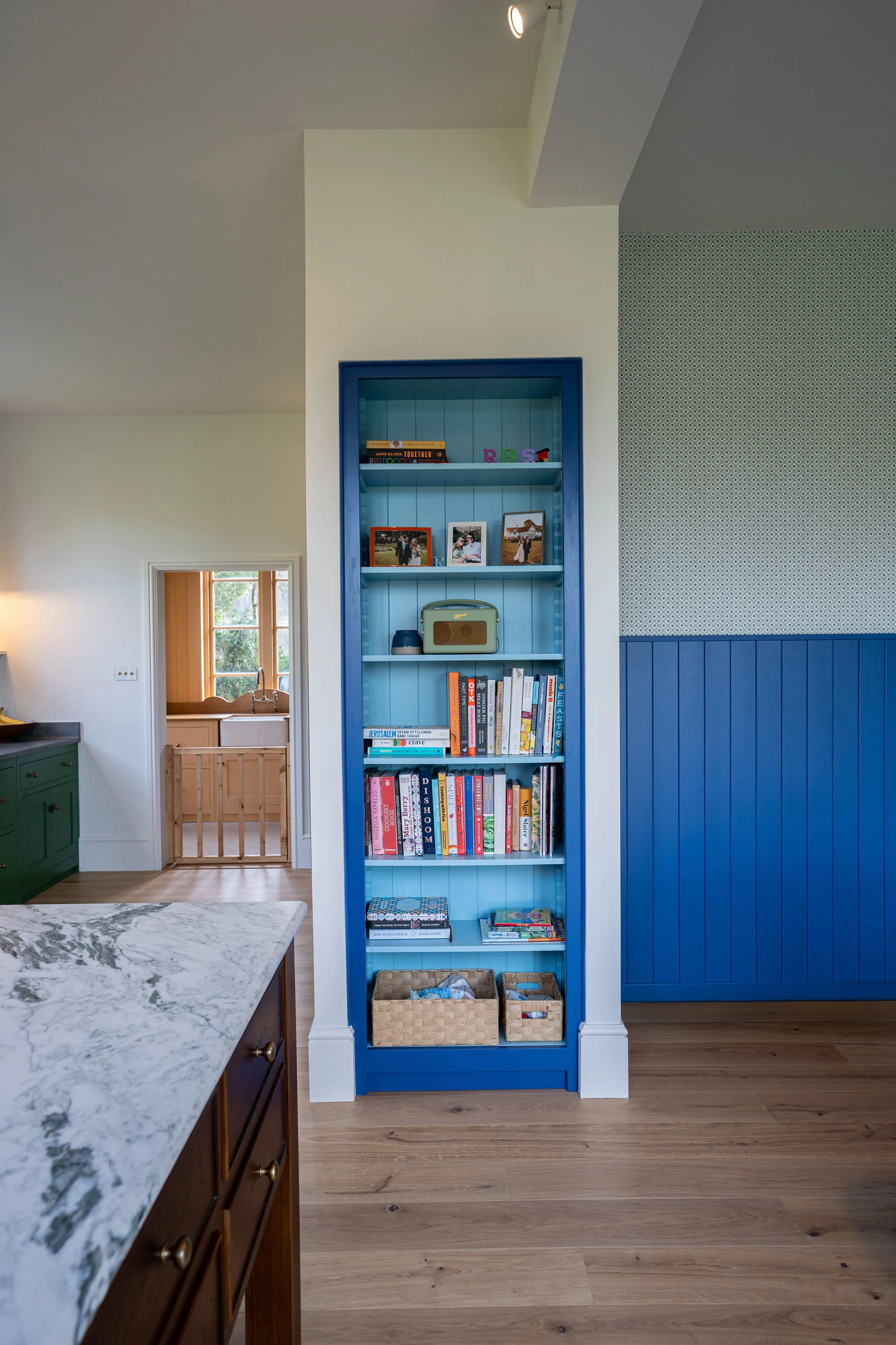 A blue bookshelf filled with books, framed photographs, a small radio, and baskets in a room with a hardwood floor and partial view of a kitchen.