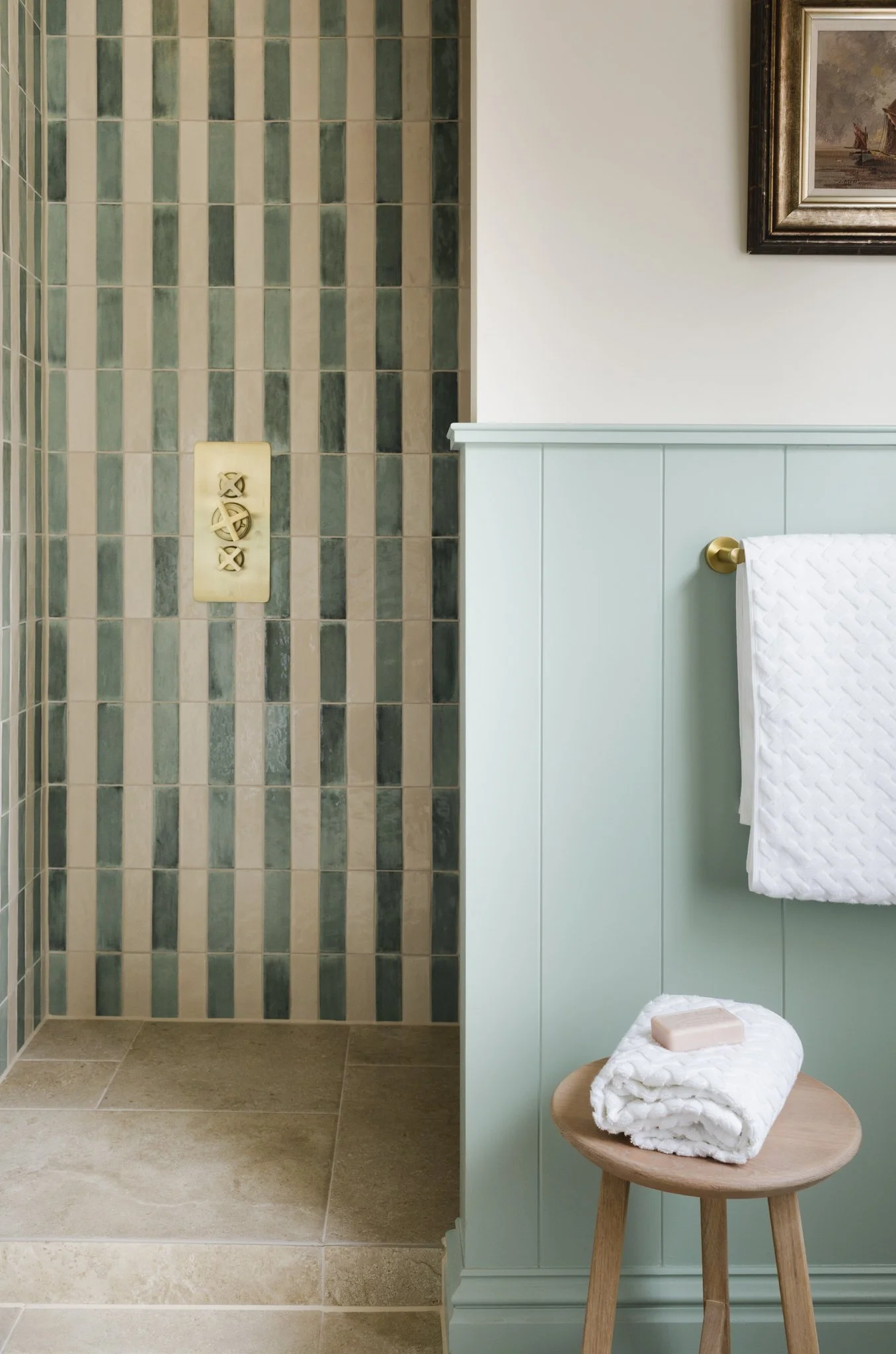 A bathroom with a tiled shower area on the left, featuring beige and green vertical tiles, and a light blue wainscot on the right with a towel rack holding a white towel. A small wooden stool with a rolled white towel and a bar of soap is in front of the wainscot. A framed painting hangs on the wall above.