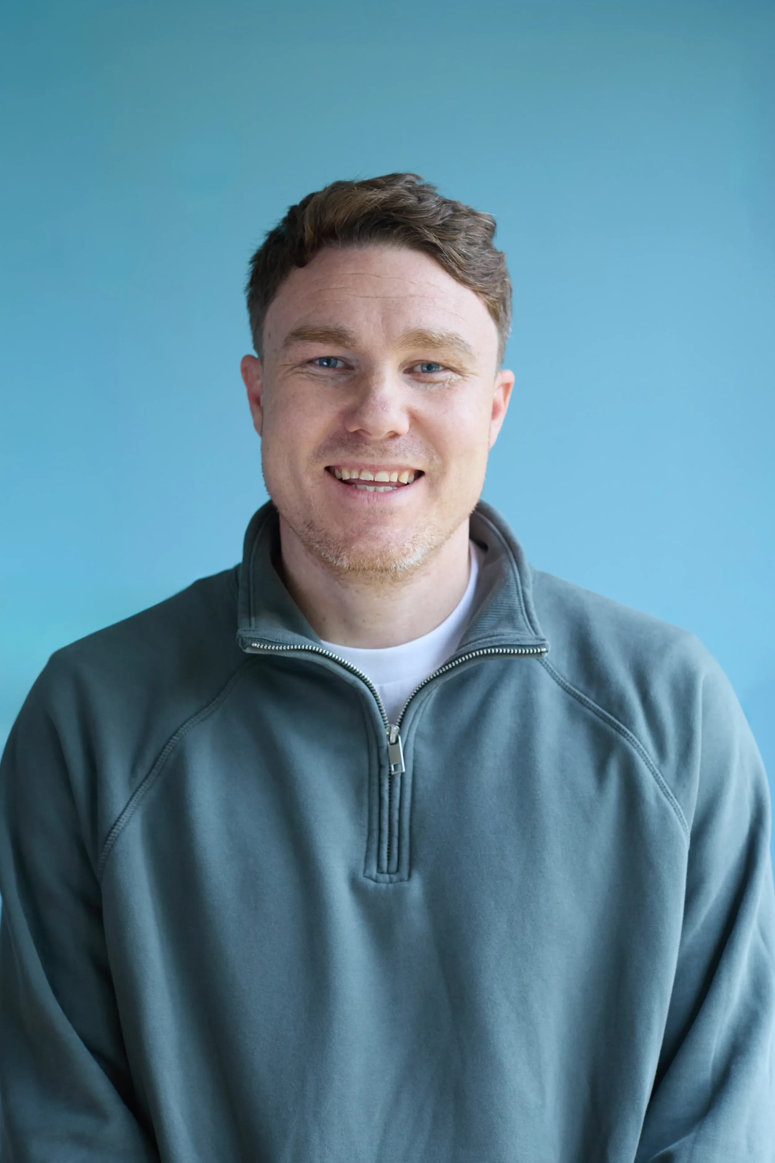 A man with light skin and brown hair, wearing a gray zip-up sweatshirt, smiling in front of a blue background.