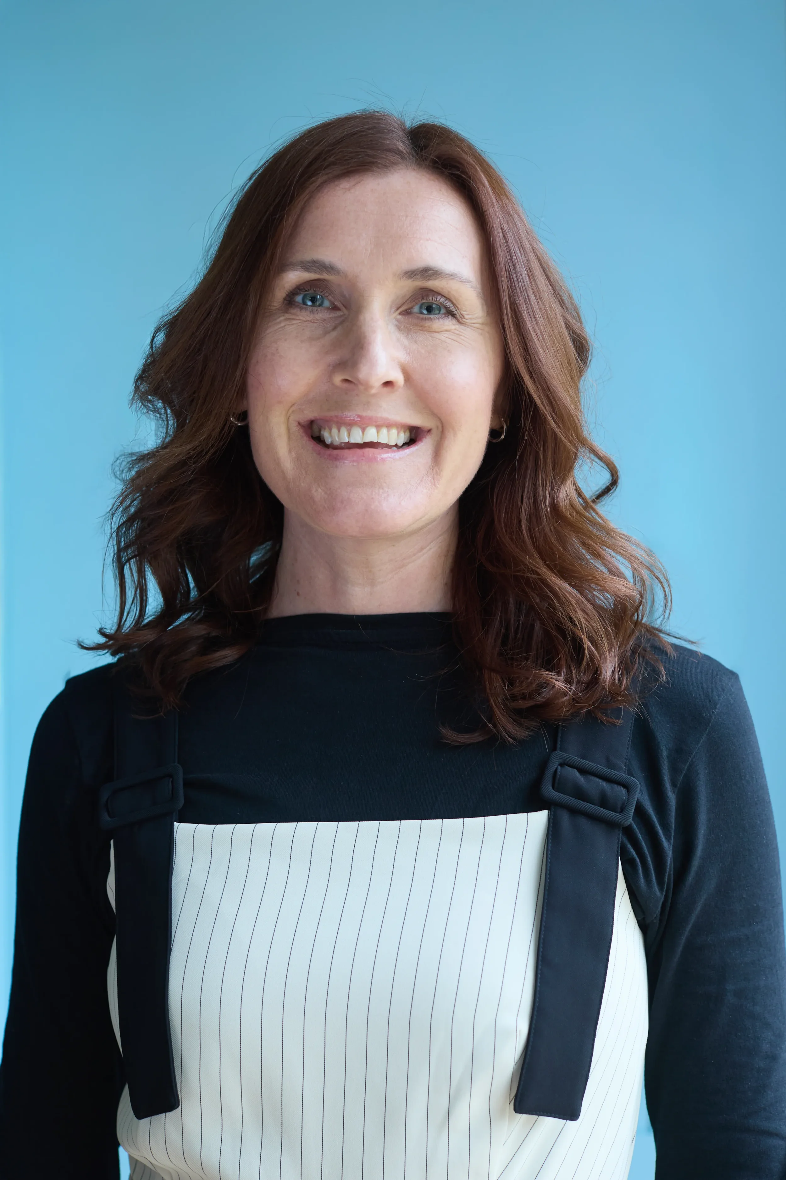 A woman with reddish-brown wavy hair smiling against a blue background. She is wearing a black jacket with a white and black striped shirt underneath and has small hoop earrings.