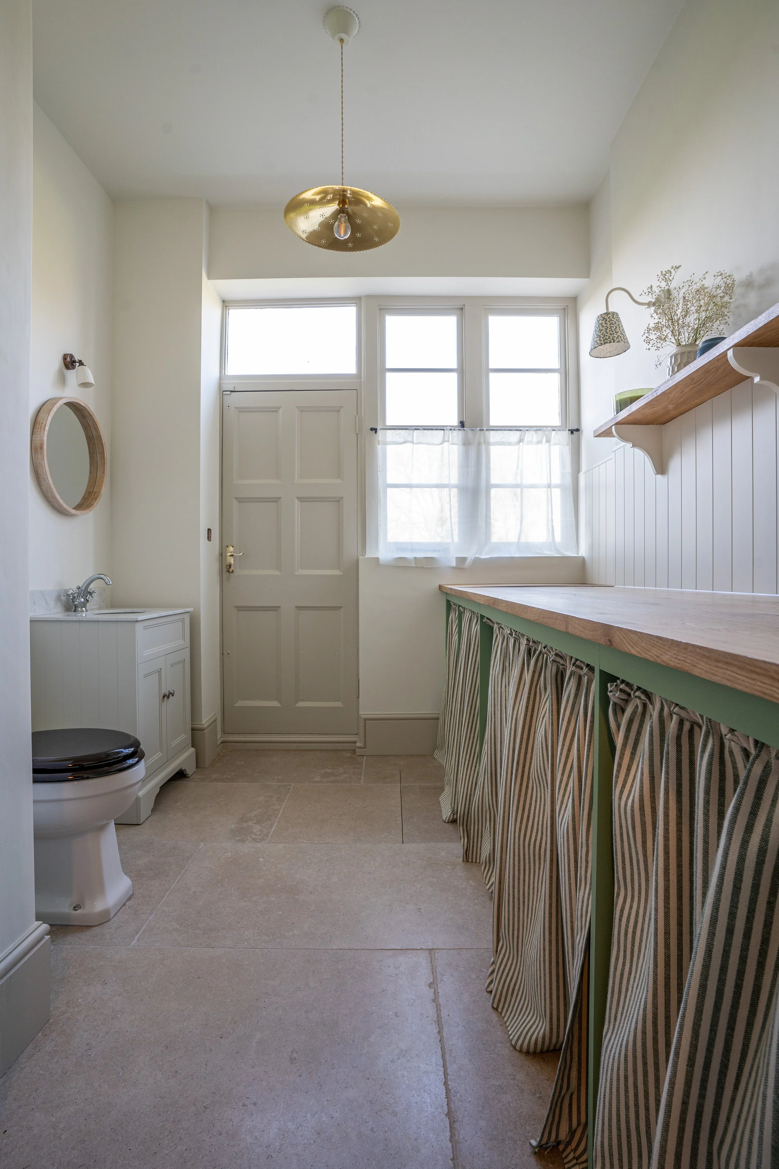 A bright minimalist bathroom with a white tiled floor, a small sink vanity, a toilet with a black seat, a window with sheer curtains, and a wooden countertop with striped curtain skirts underneath. A gold ceiling light and a mirror hang on the wall.
