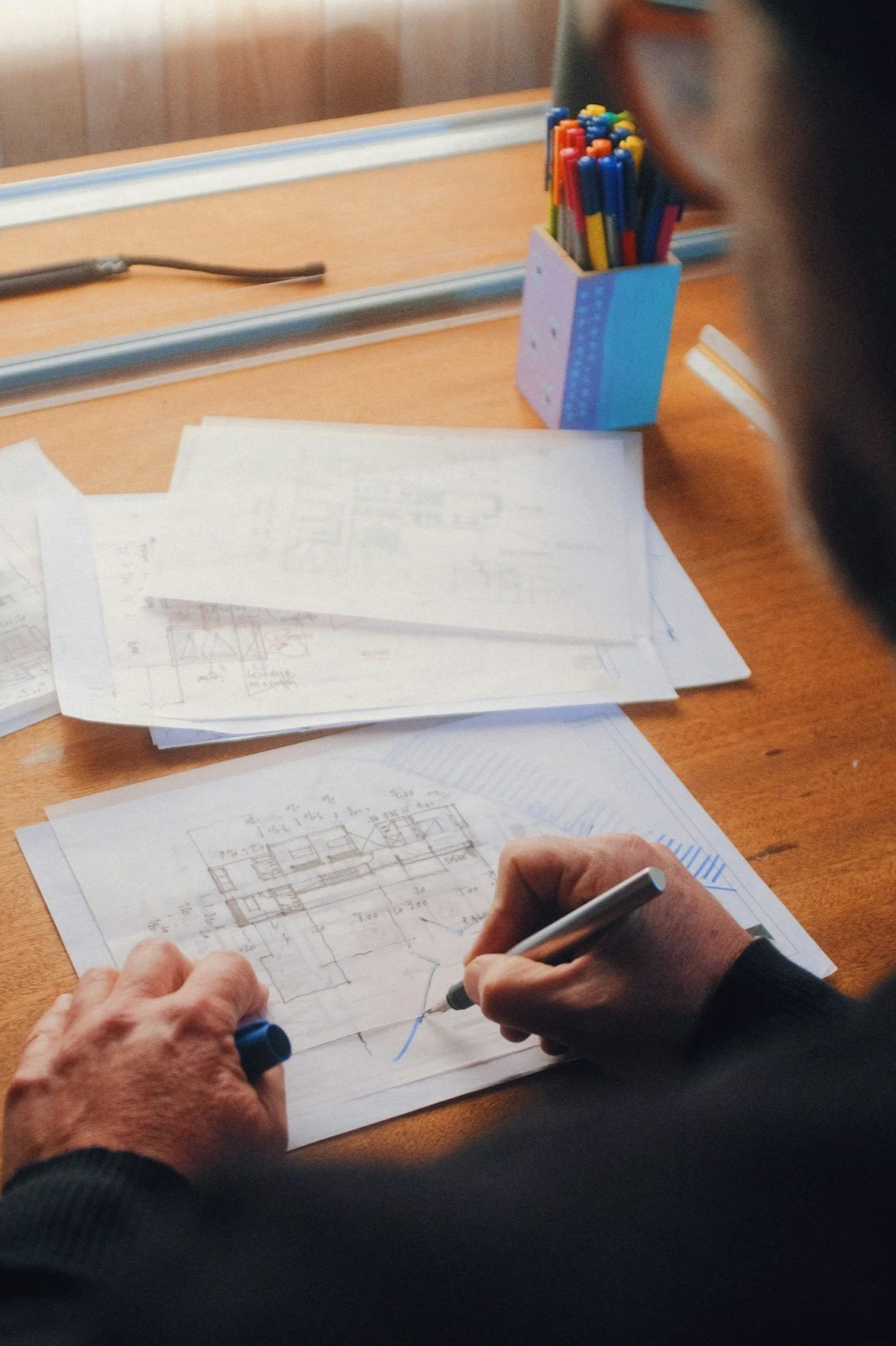 Someone sketching architectural plans on paper at a wooden desk, surrounded by additional papers and a container of colored pens.
